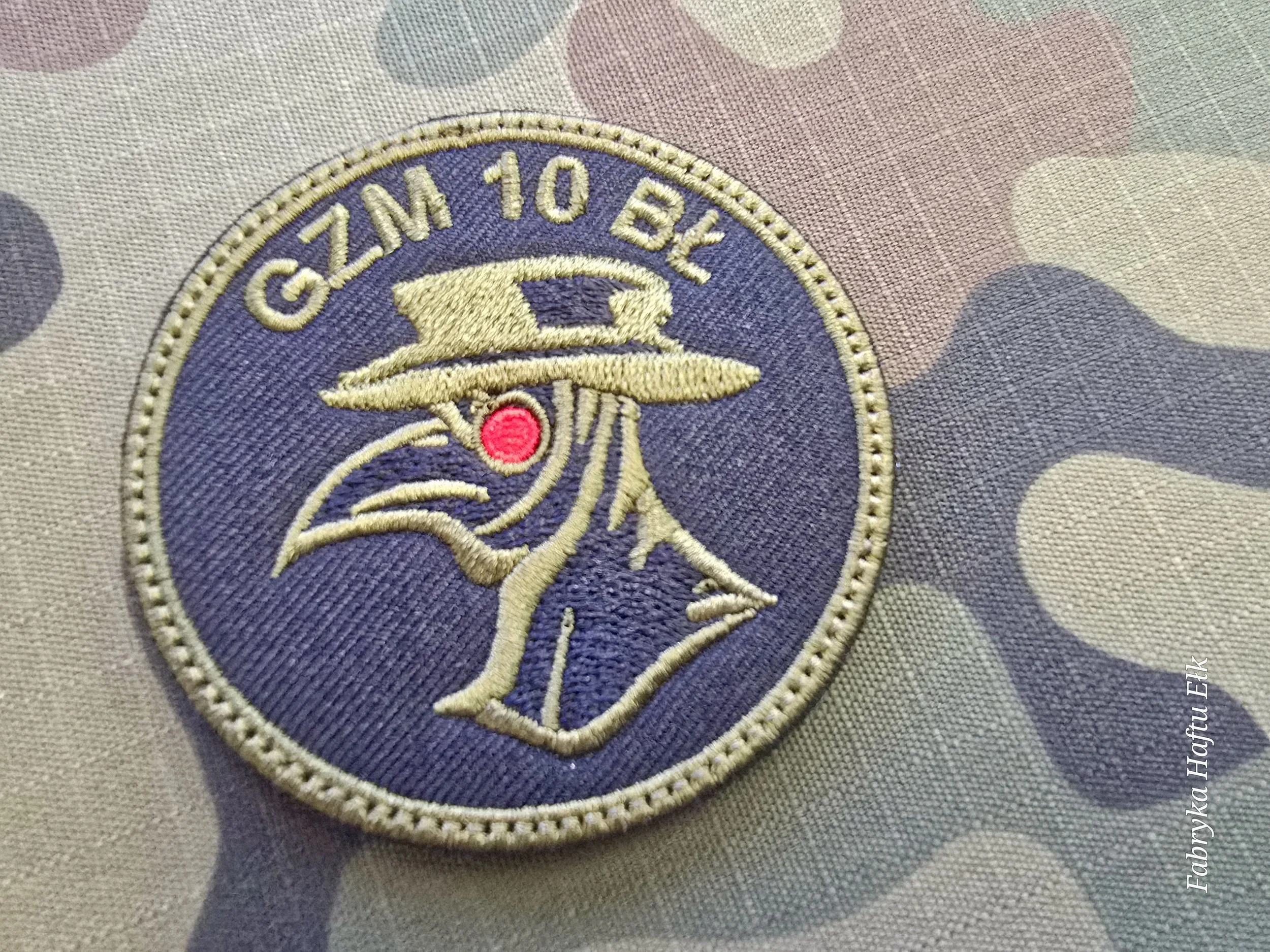 Okrągła naszywka GZM 10 BL z postacią w masce z długim dziobem i czerwonym okiem, na tle kamuflażu