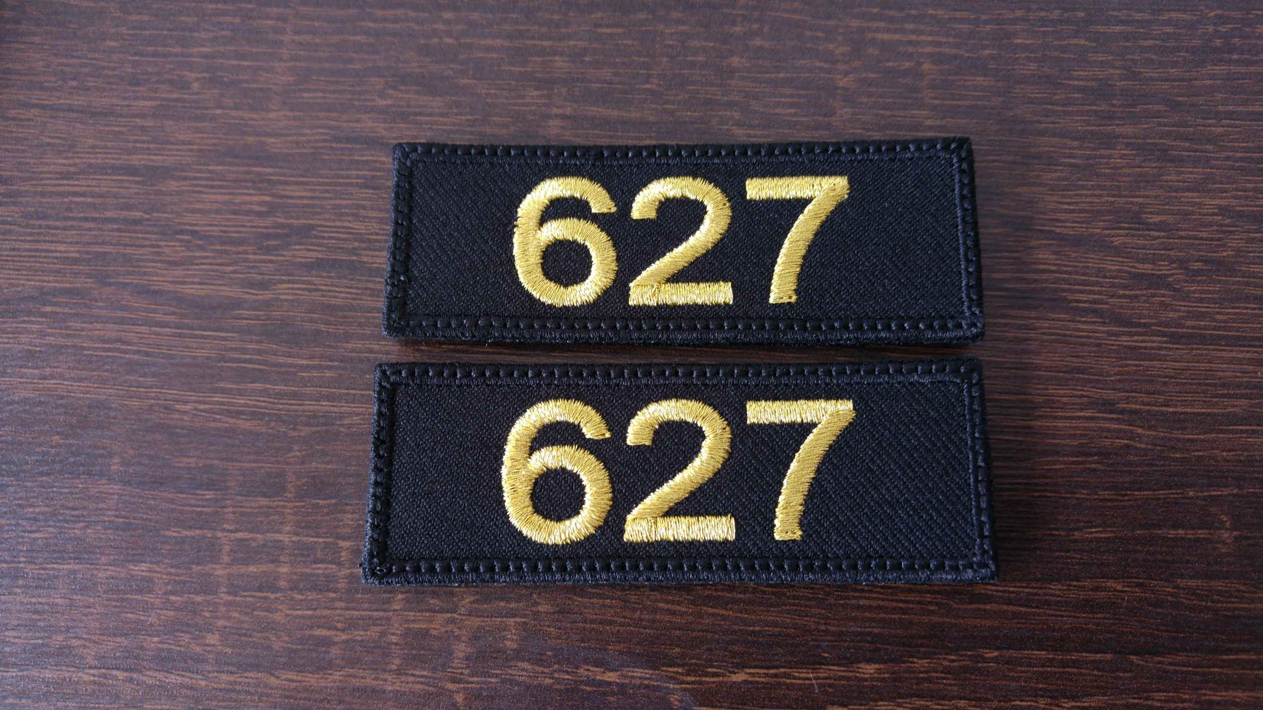 Two black fabric patches with the number 627 embroidered in gold, placed on a wooden surface.