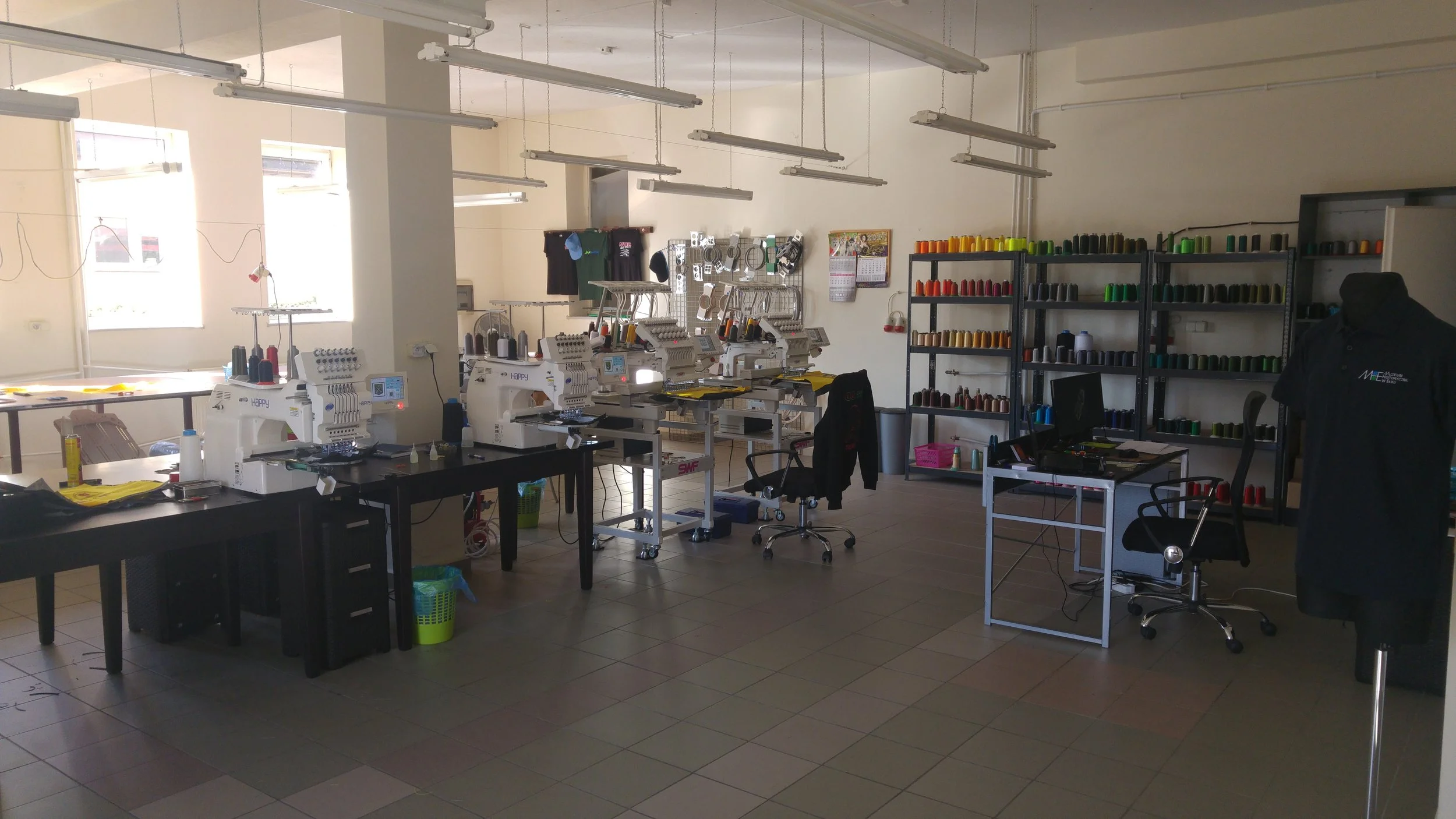 A sewing workshop with multiple sewing machines on black tables, shelves filled with colorful spools of thread, and large windows allowing natural light, with chairs, a dress form, and various sewing tools and materials visible.