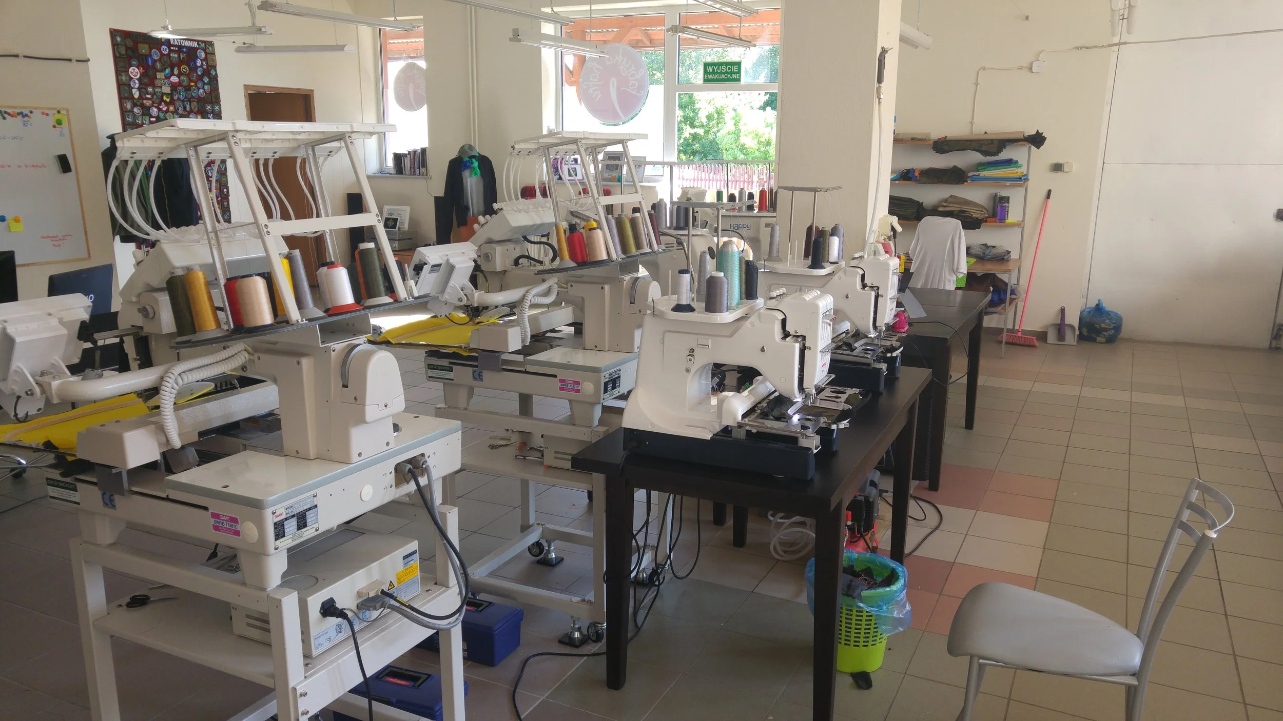A room filled with multiple sewing machines, spools of thread, and fabric on shelves. There are chairs, a whiteboard, and a door with a green exit sign in a well-lit environment.