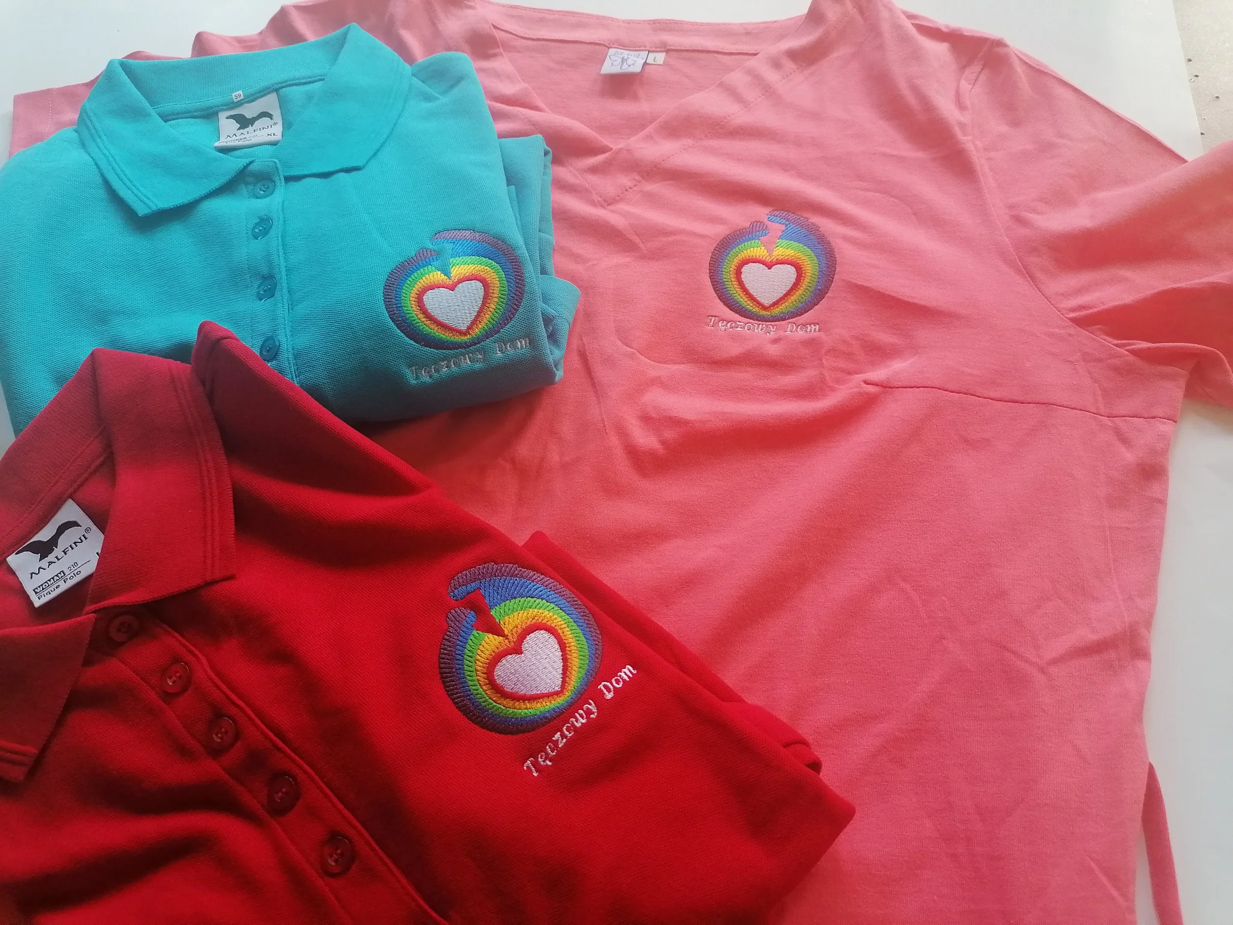 A collection of clothing items including a light blue embroidered polo shirt and a pink shirt, both with rainbow heart and flame designs, along with a red garment, all laid out on a white surface.