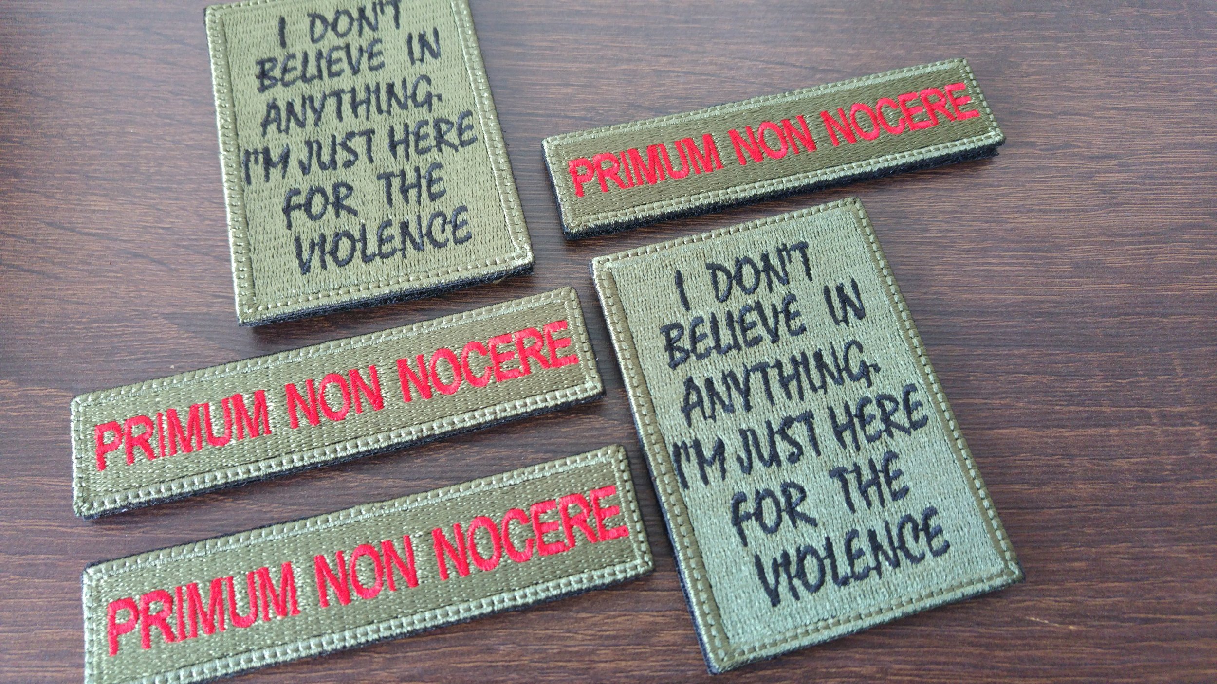 Several fabric patches with embroidered text, including phrases 'I don't believe in anything my just here for the violence' and 'Primum Non Nocere' in red and black thread, placed on a wooden surface.