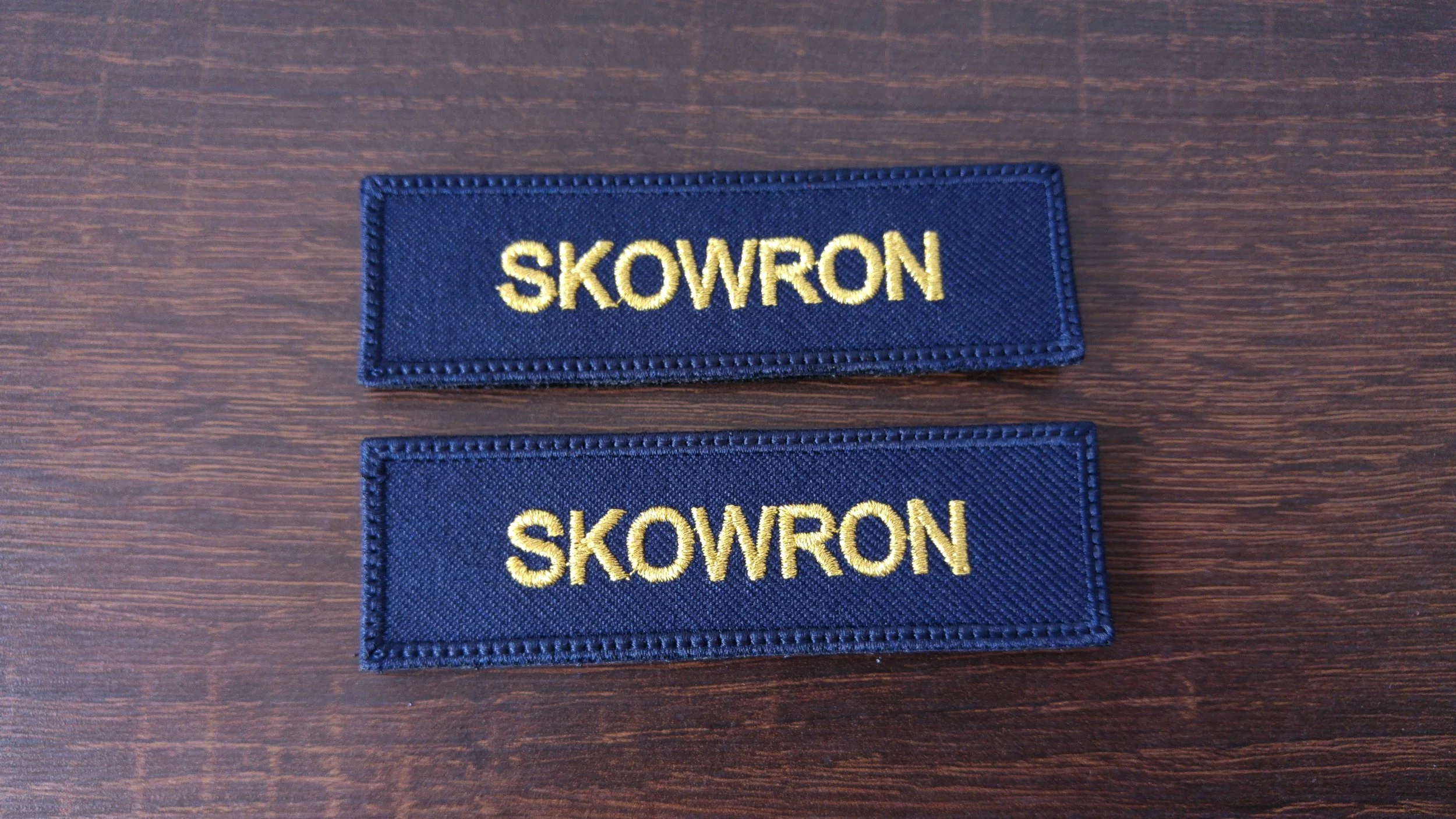 Two dark blue embroidered patches on a wooden surface, each with the word "SKOWRON" in yellow lettering.