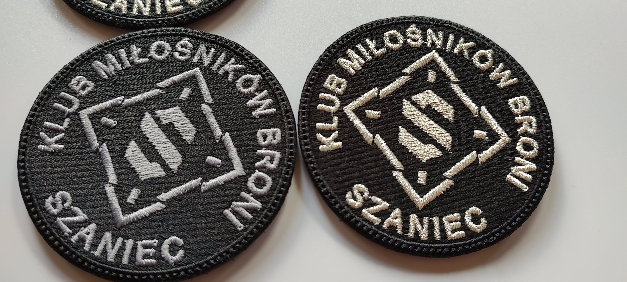 Czarne naszywki Klubu Miłośników Broni Szaniec z jasnym haftowanym symbolem i napisami