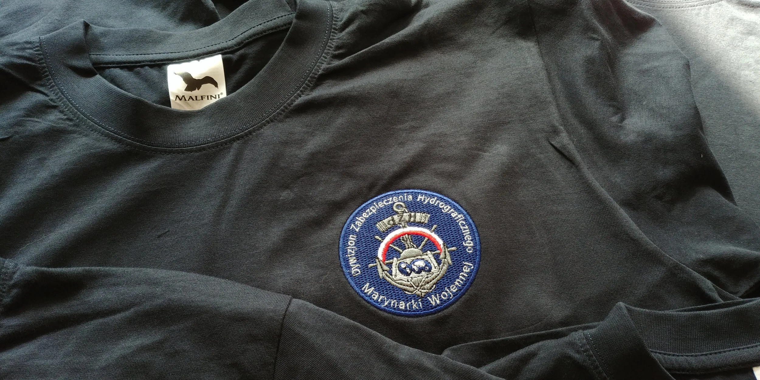 A black T-shirt with a round badge featuring Polish text and an emblem. The shirt has a tag from Malfini. The badge contains a nautical or maritime symbol.