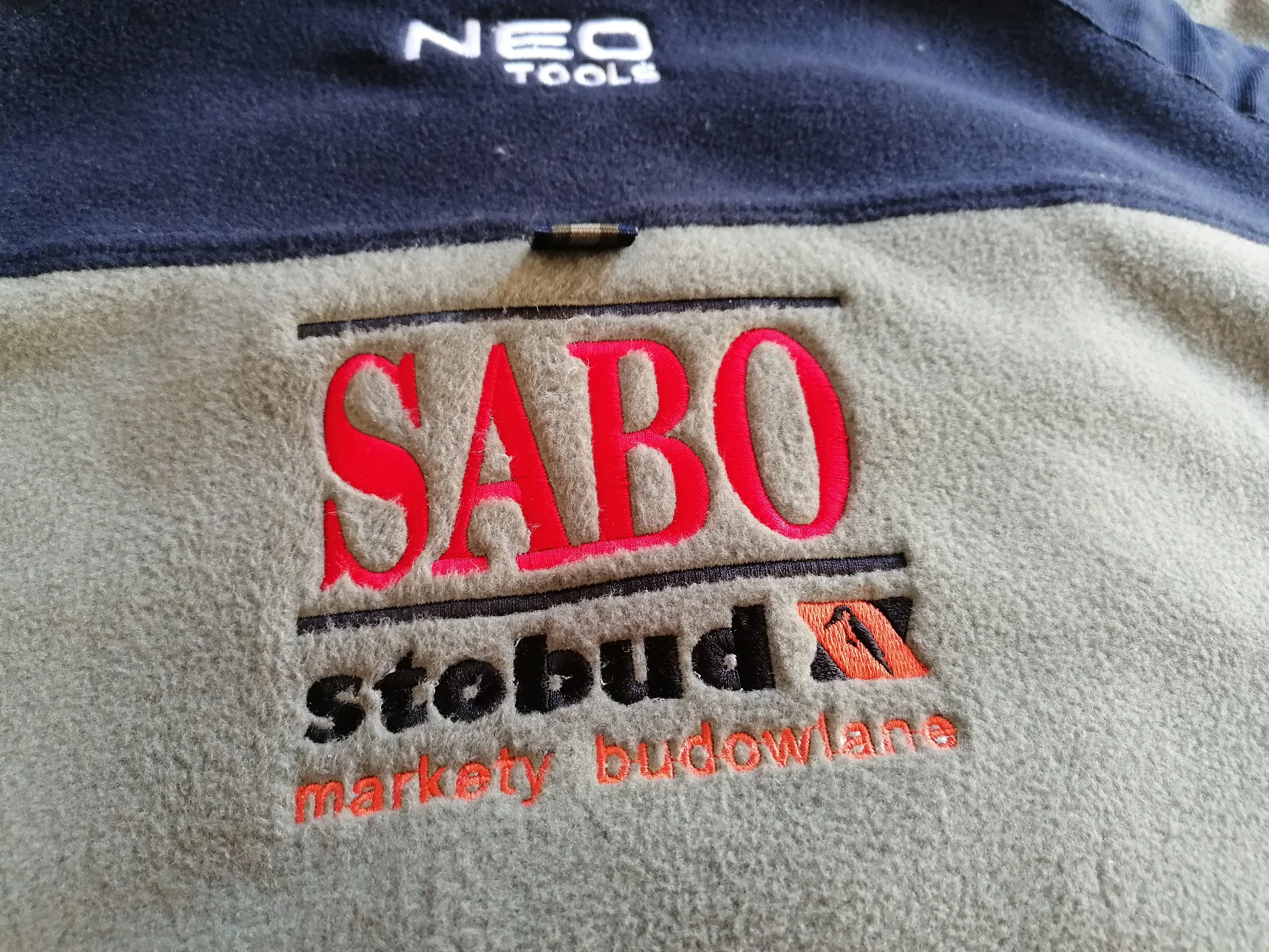 Close-up of embroidered patch on a fleece jacket, showing the word 'SABO' in red letters, and portions of other embroidered words and logos including 'NEO TOOLS' at the top.