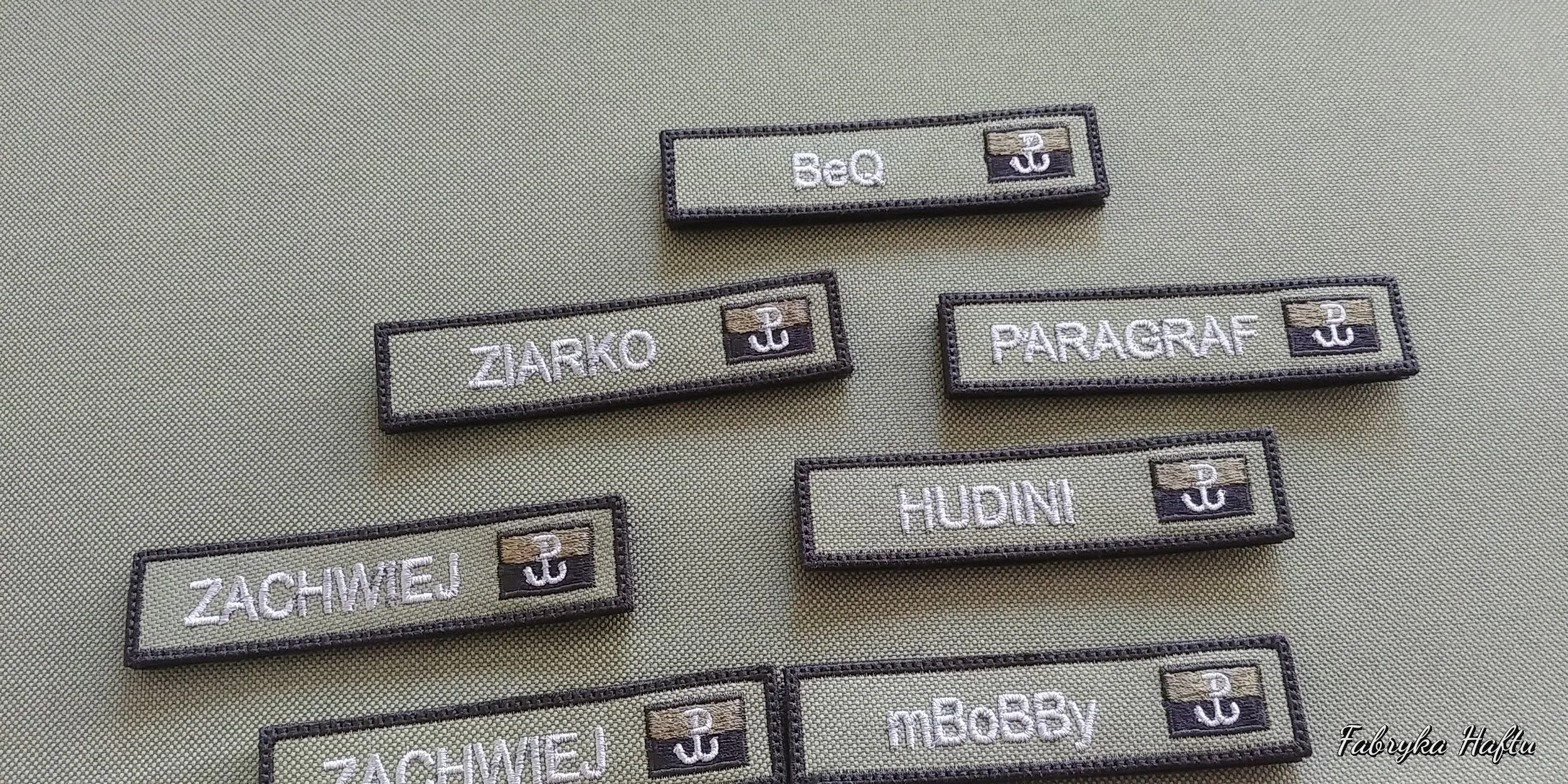 Several fabric name tags with black borders on a green surface. The tags have various names embroidered in white, such as BeQ, ZACHMEJ, PARAGRAF, HUDIN, and MEBBXY, each accompanied by a small emblem of an anchor.