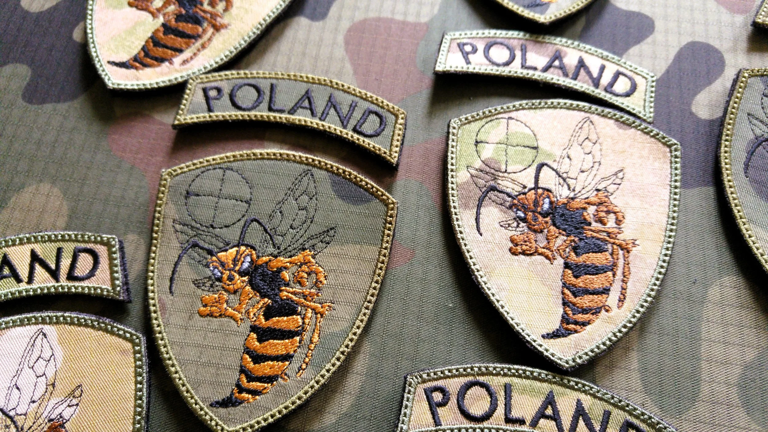 Multiple embroidered patches featuring the word 'POLAND' and a bee logo on a camouflage fabric background.