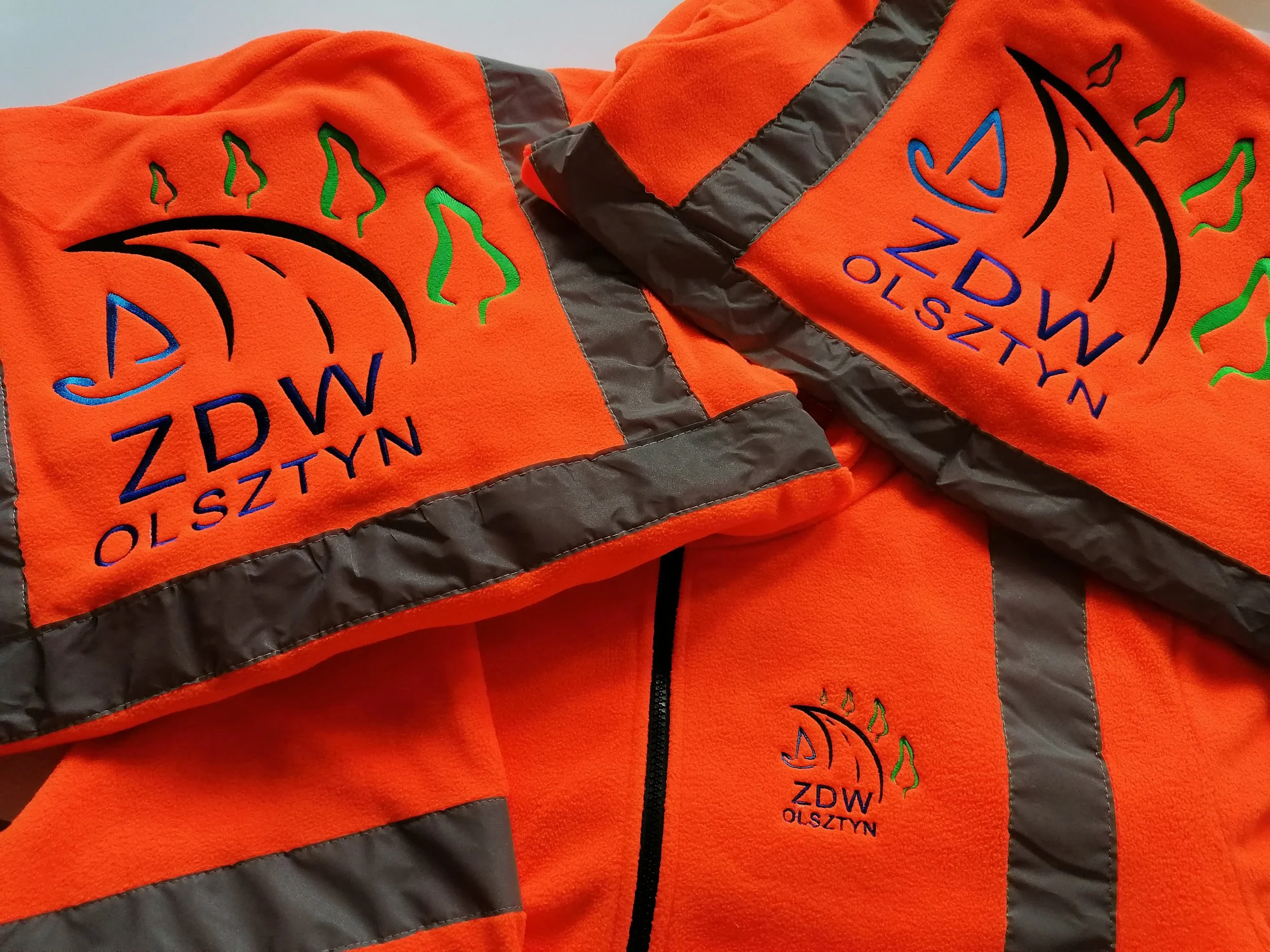 Close-up of bright orange fleece jacket with black reflective stripes and embroidered logo with green, blue, and black outline of a pumpkin and the words "MALISZYN ZDZWOY" in blue.