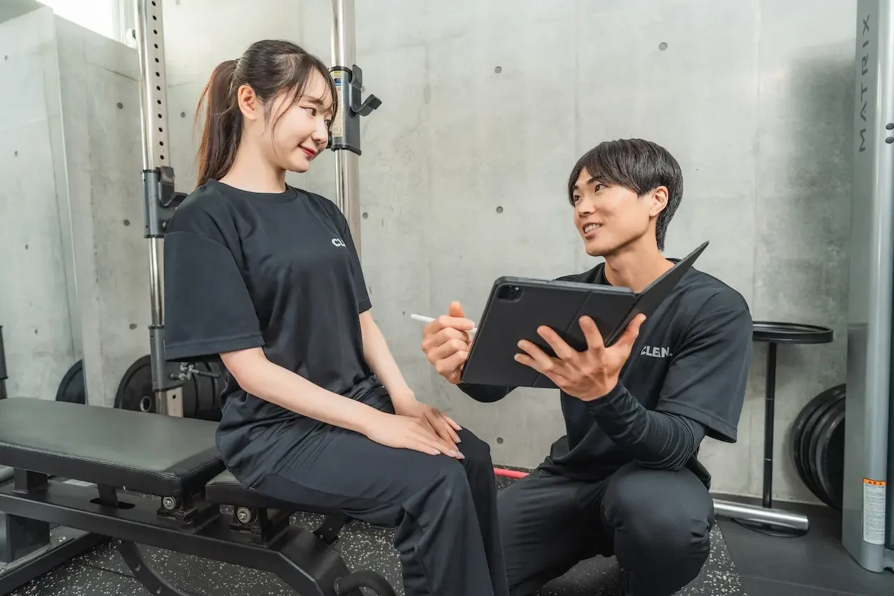 日吉のジム「CLEAN.」にて、トレーニングのインターバル中に行われるパーソナルストレッチ。休憩時間も無駄にせず、トレーナーが筋肉の可動域を広げる調整を行うことで、怪我を防ぎながら効率的にボディメイクを促進している指導風景。
