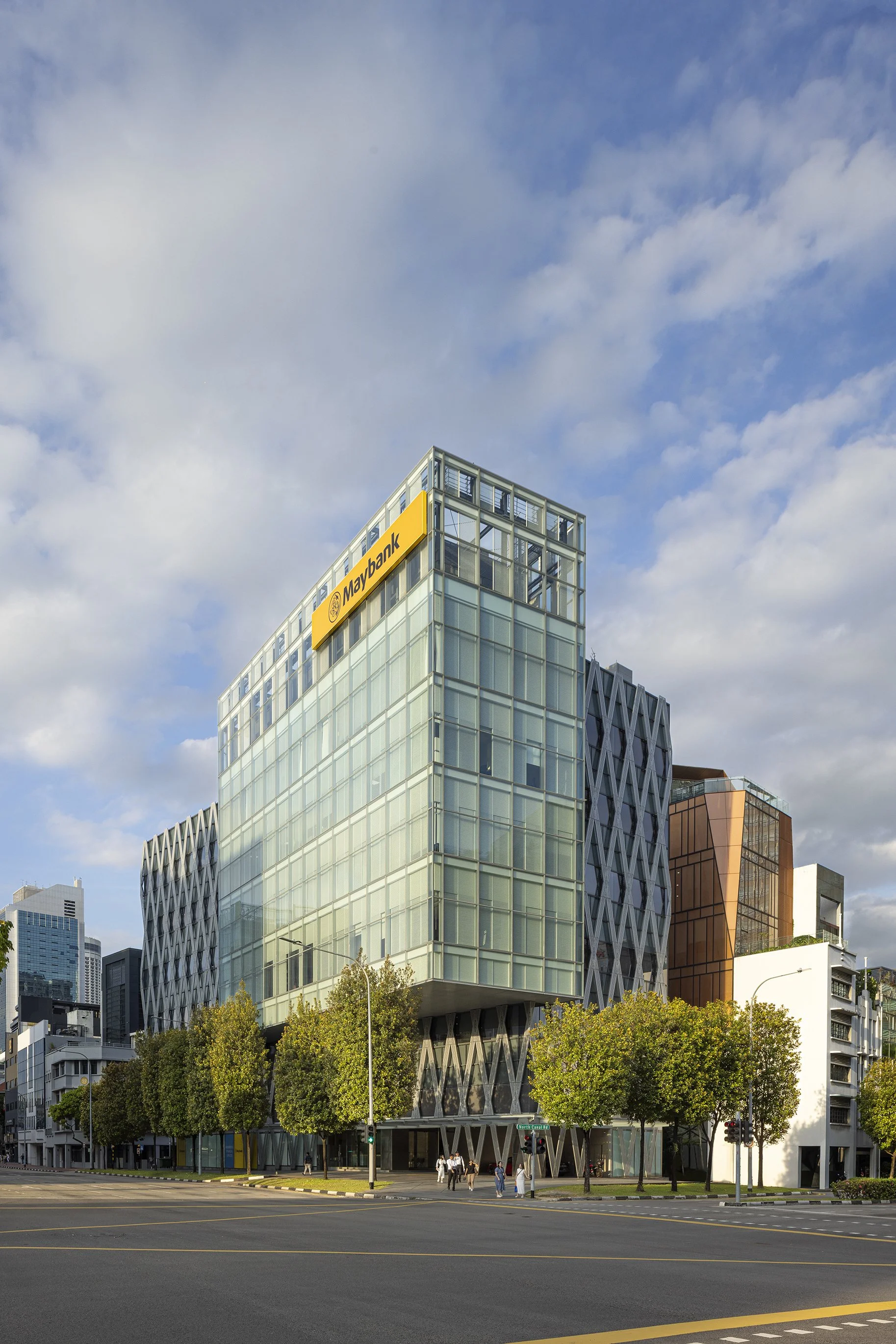 Modern office building with Maybank sign, glass facade, and trees in front, under a partly cloudy sky