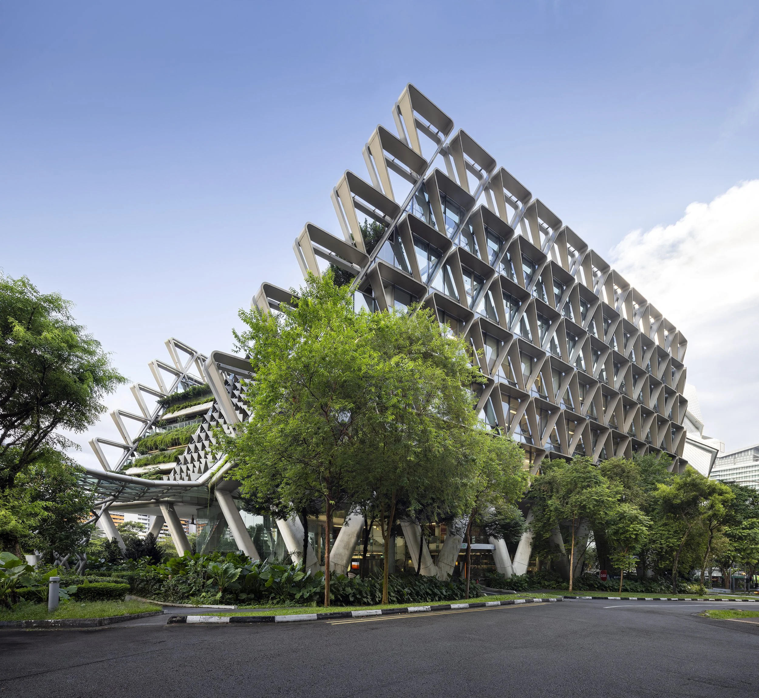 Modern multi-story building with an intricate facade design, surrounded by greenery and trees, under a clear sky.