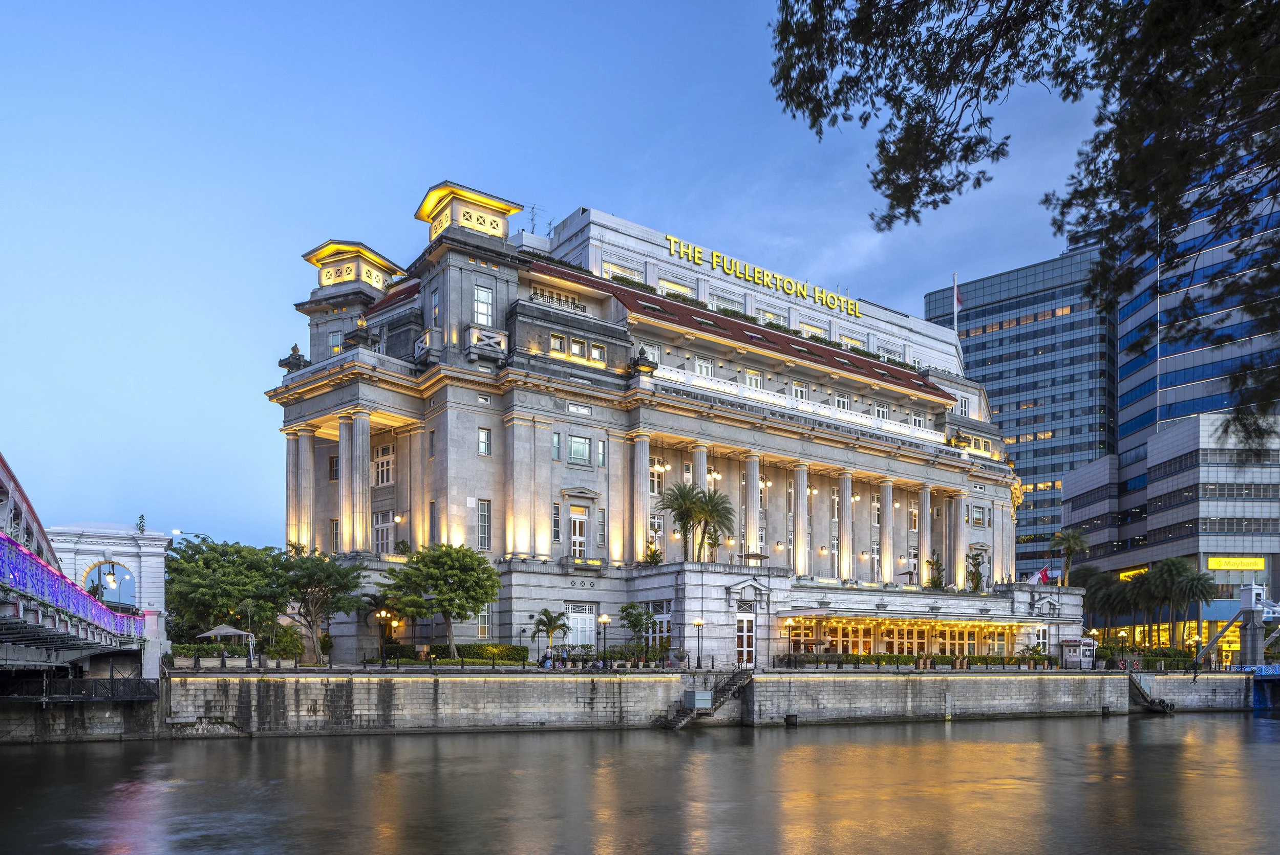 Exterior of The Fullerton Hotel in Singapore during dusk with city skyline behind and river in foreground.