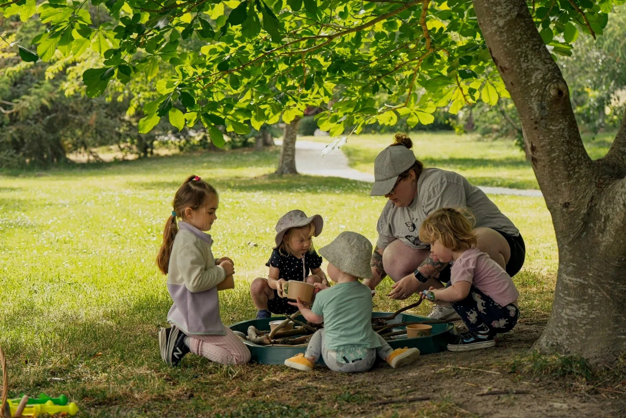 A group of children and a woman playing outdoors under a large tree with green leaves, sitting around a small sandbox with sticks and toys.