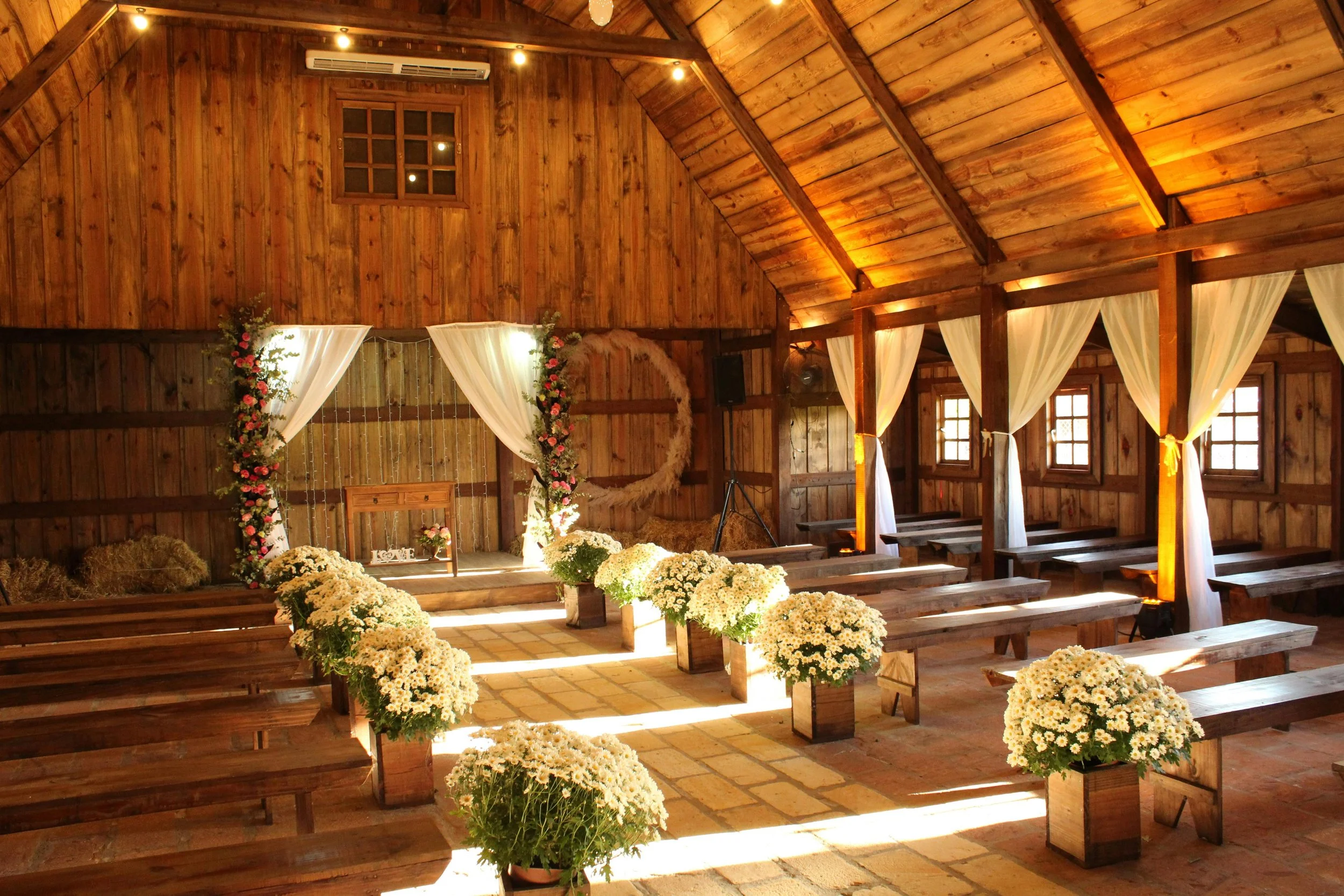 Rustic barn wedding ceremony space with wooden pews, white draping, floral arch, and clusters of white flowers lining the aisle