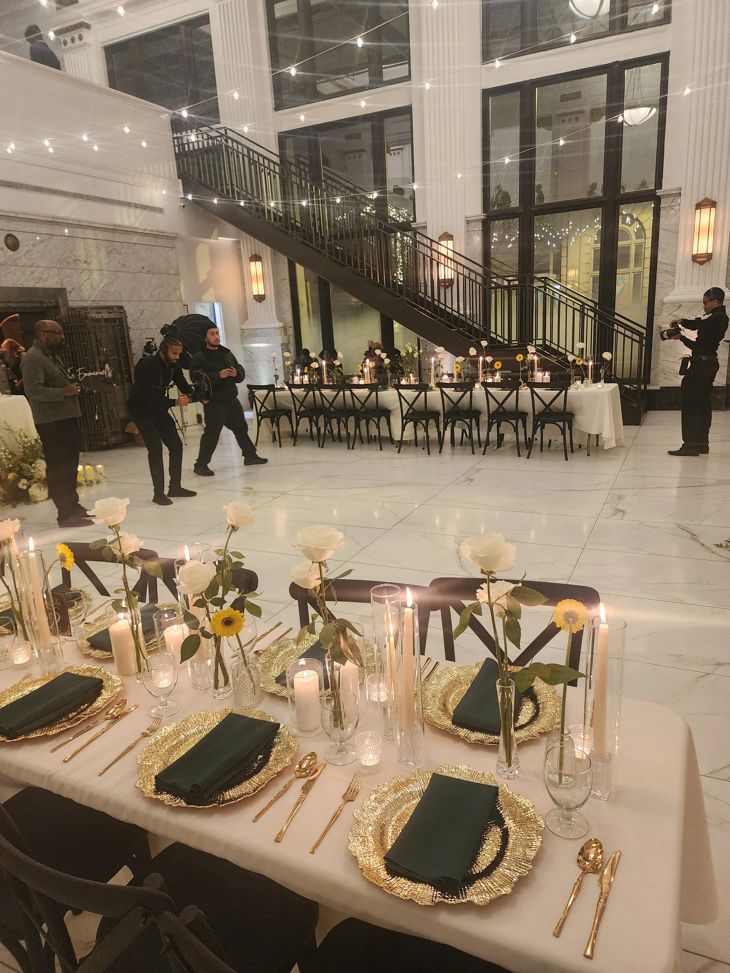 Elegantly set wedding reception tables with gold charger plates, green napkins, white roses, sunflowers, and candles inside a grand marble atrium with string lights and a staircase