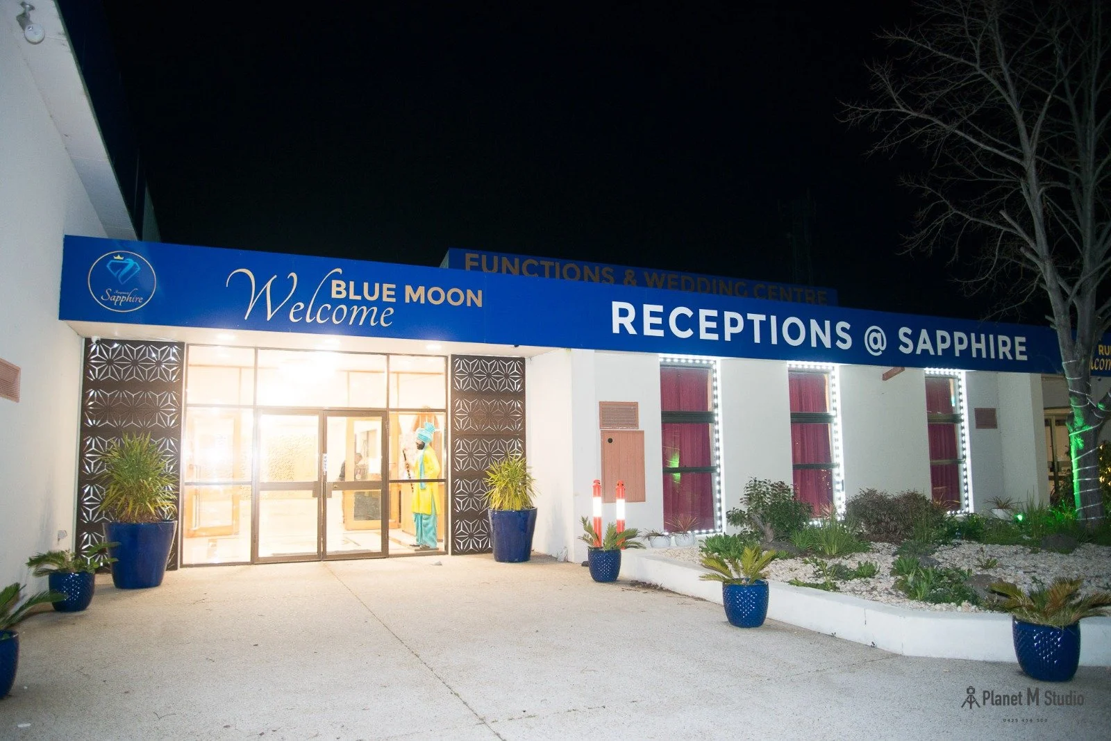 Night view of the reception entrance to the Sapphire Blue Moon wedding and event center, with potted plants and illuminated windows.