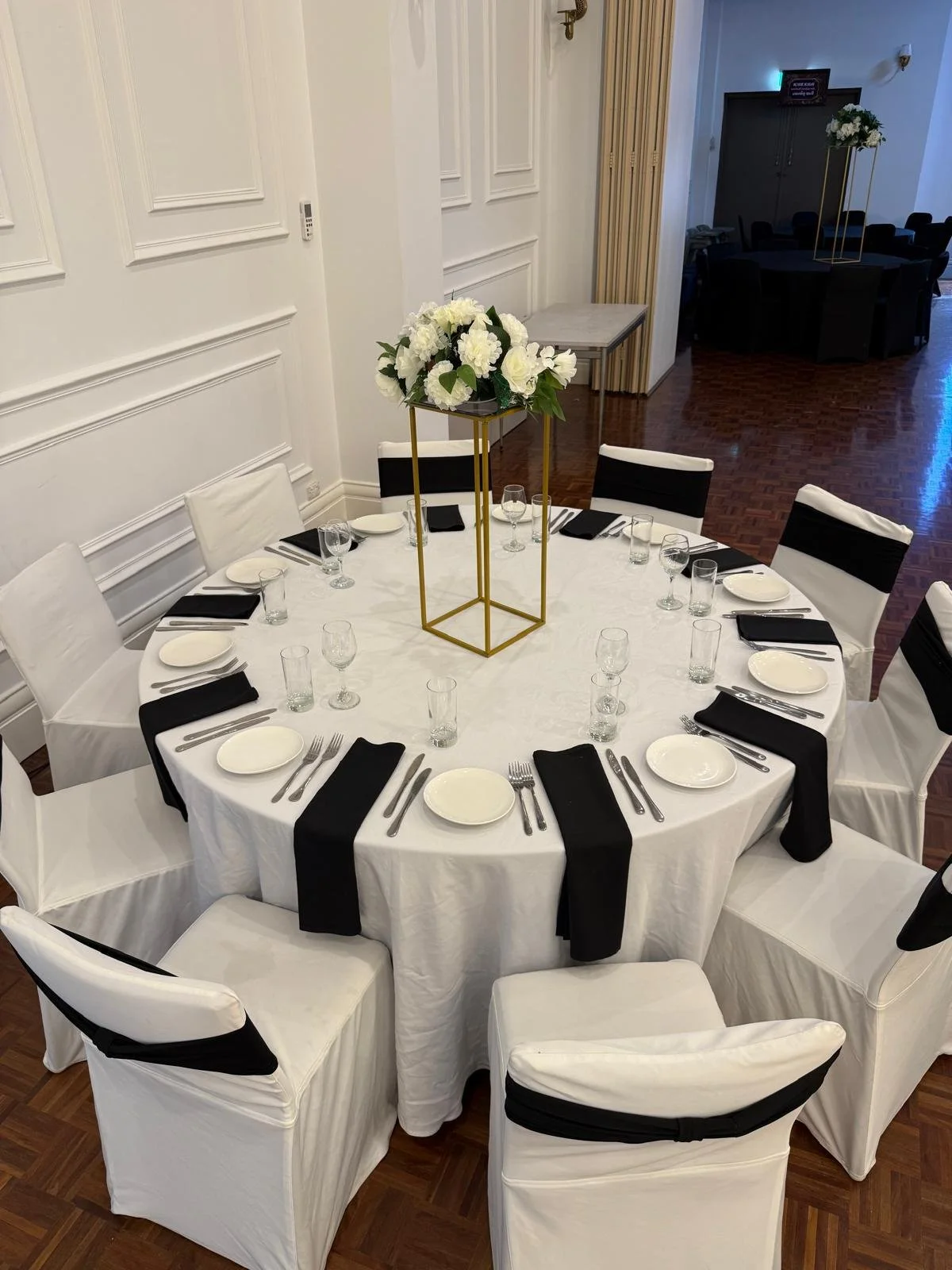 A round banquet table set with white tablecloth, white chairs with black sashes, black napkins, white plates, silverware, water glasses, and a tall gold stand with a white floral arrangement in the center.
