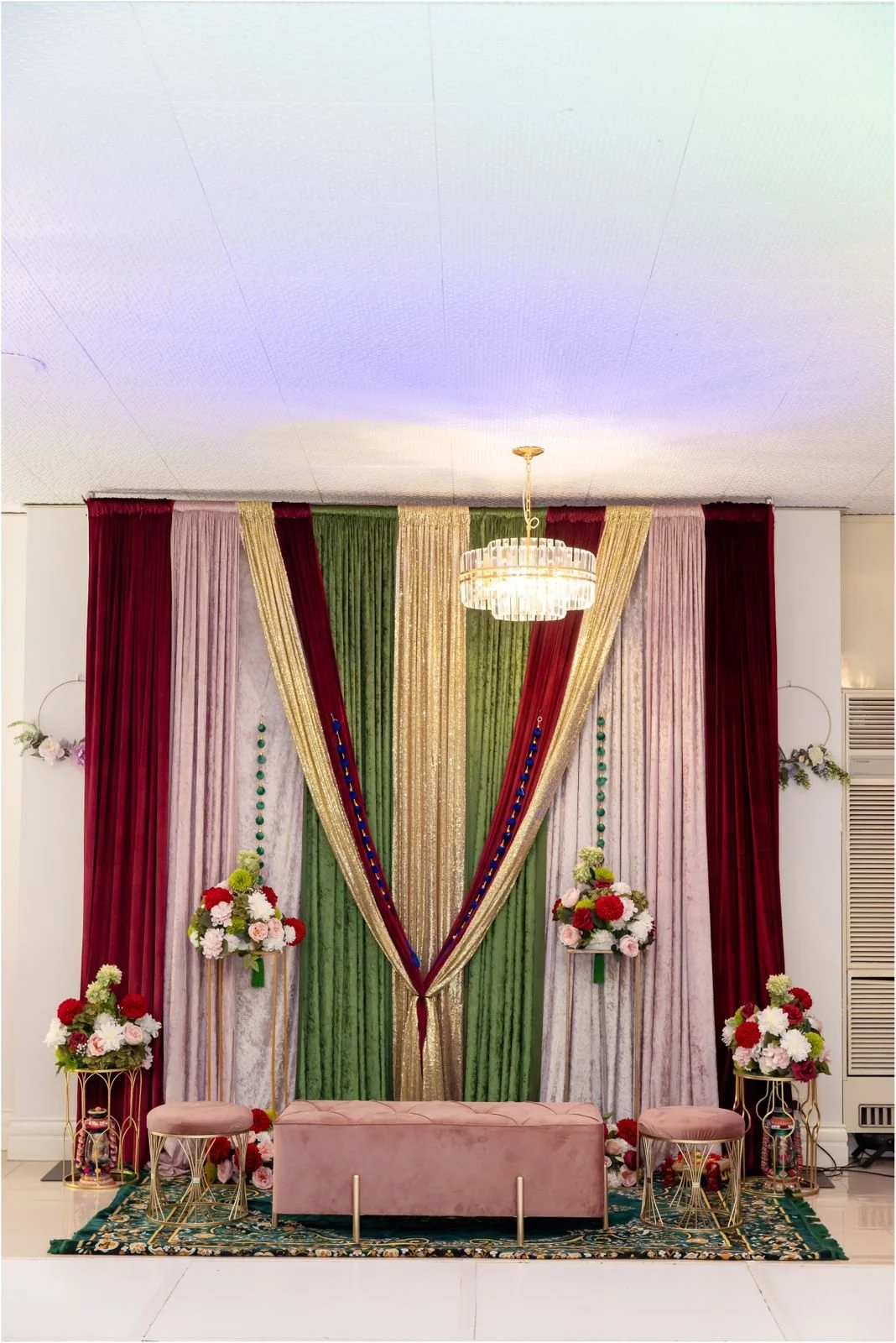 Decorative backdrop with colorful curtains and floral arrangements, pink velvet bench and stools in front, chandelier hanging from ceiling.