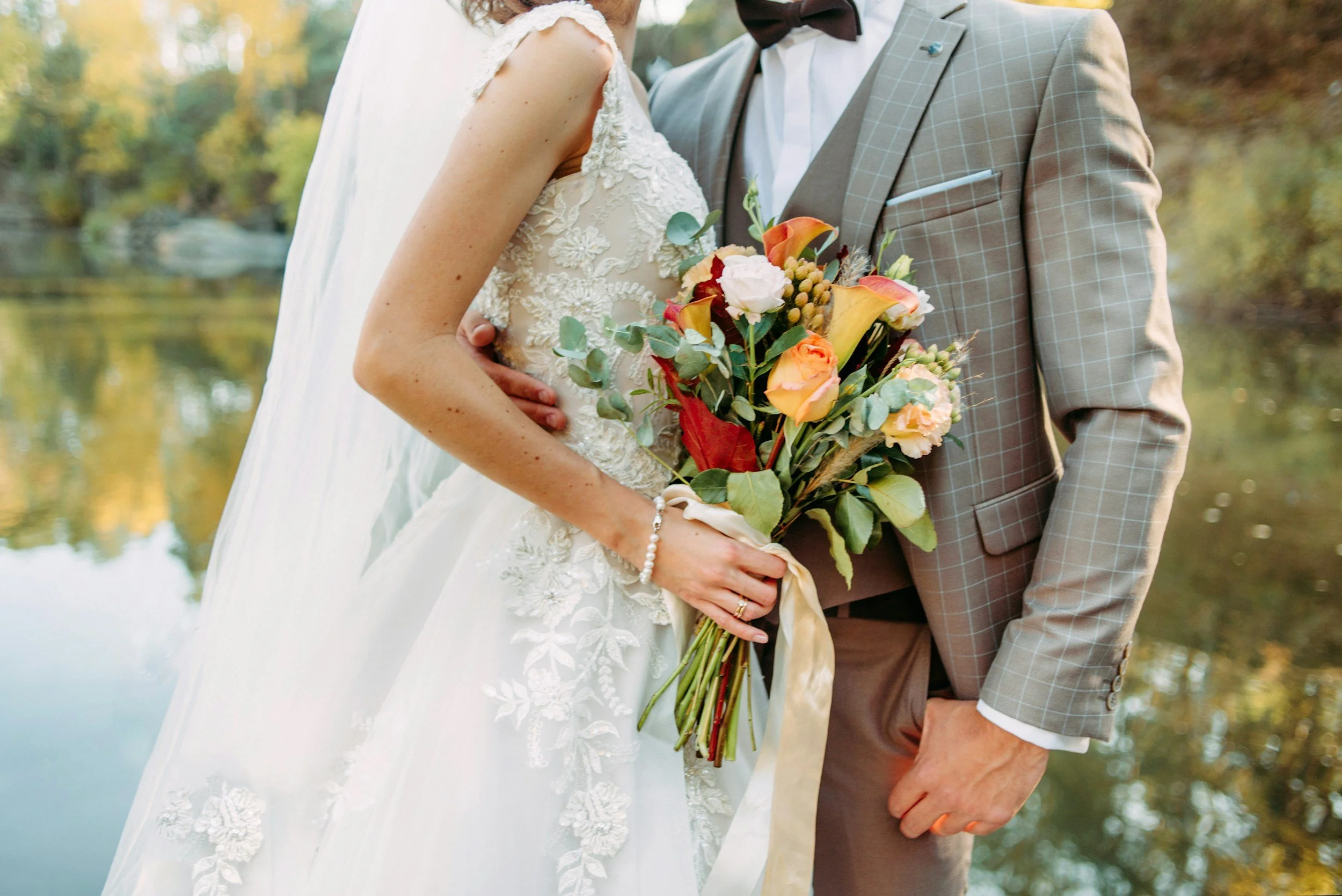 Close-up of a bride and groom holding a bouquet of flowers by a lake with trees in the background.