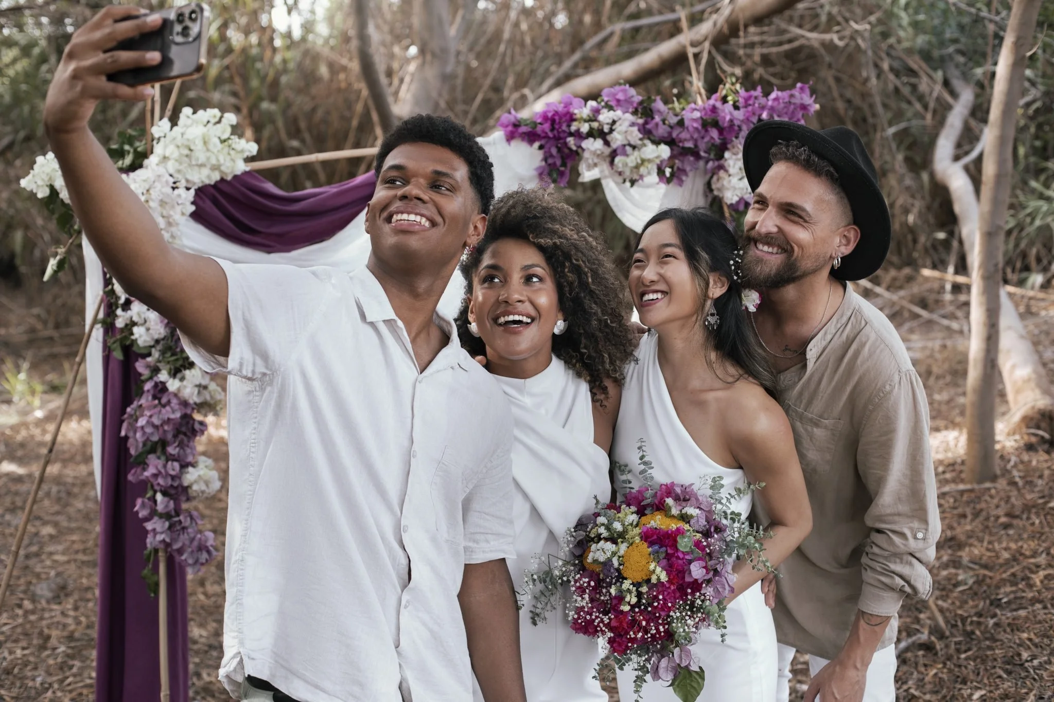 Group of four diverse people taking a selfie at a wedding ceremony outdoors, with a decorated floral arch behind them.