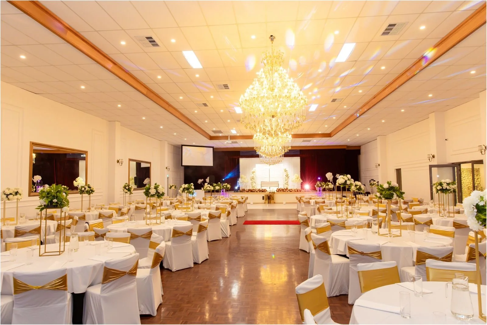 Elegant banquet hall decorated for a wedding or special event with white tablecloths, gold accents, floral centerpieces, chandeliers, and a stage at the front.