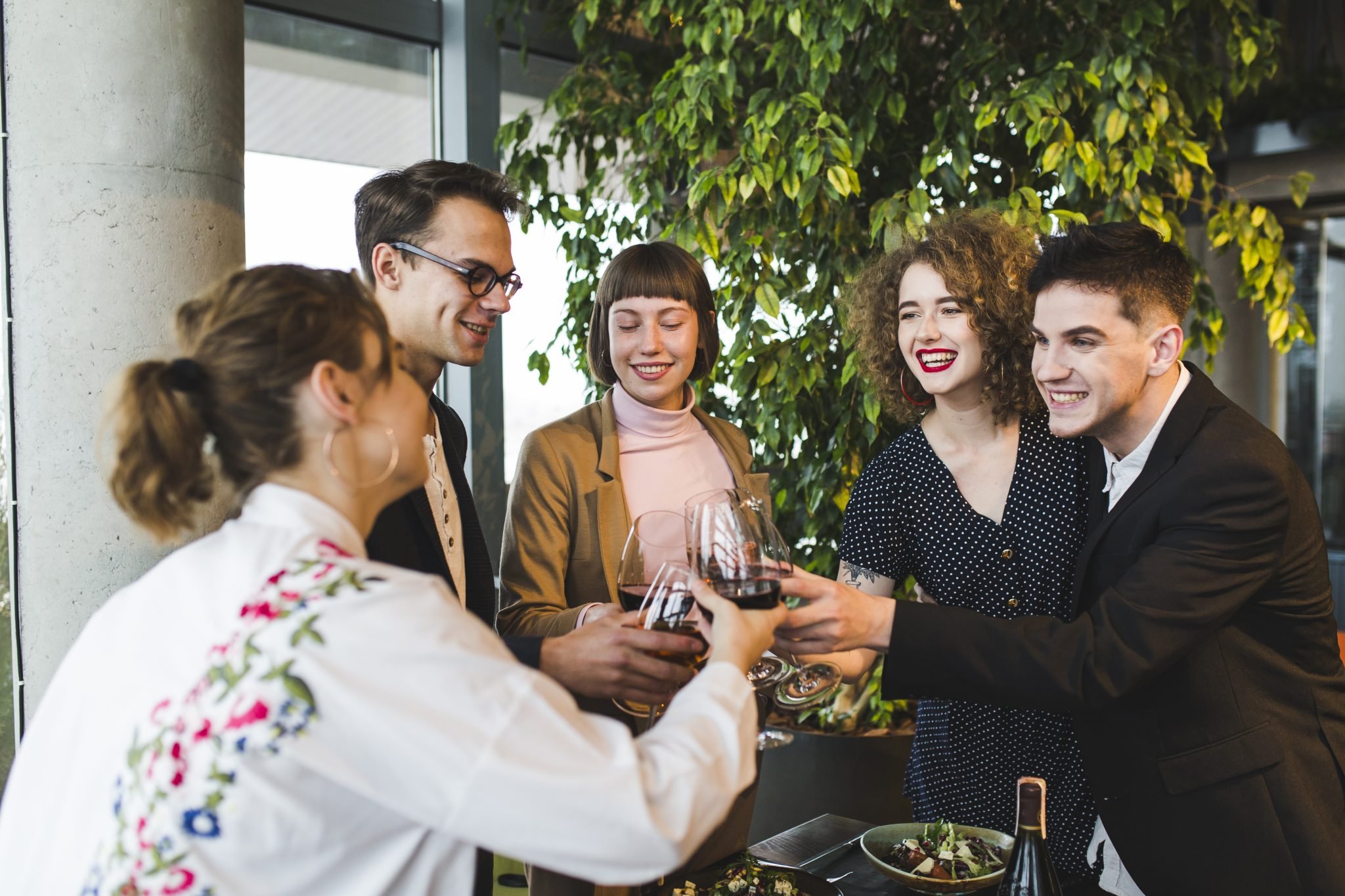 Group of five young adults enjoying wine and food in a modern, well-lit restaurant with large windows and greenery.