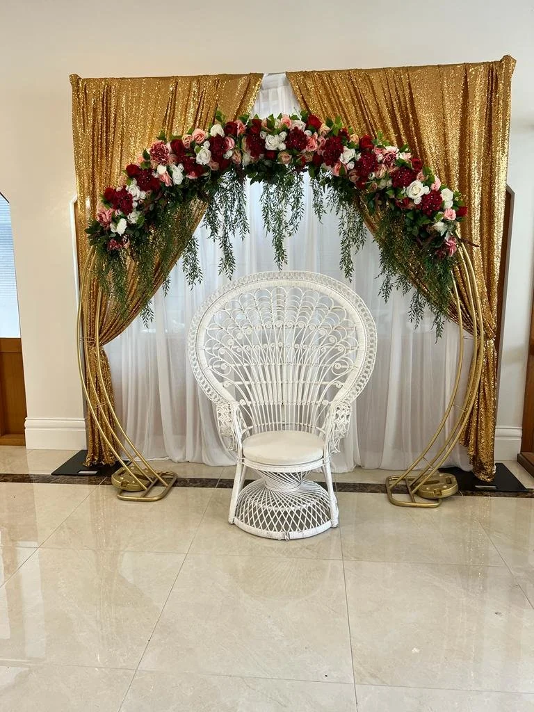 Gold sequin curtains with a floral arch and white peacock chair in front, set up for a celebration or event.