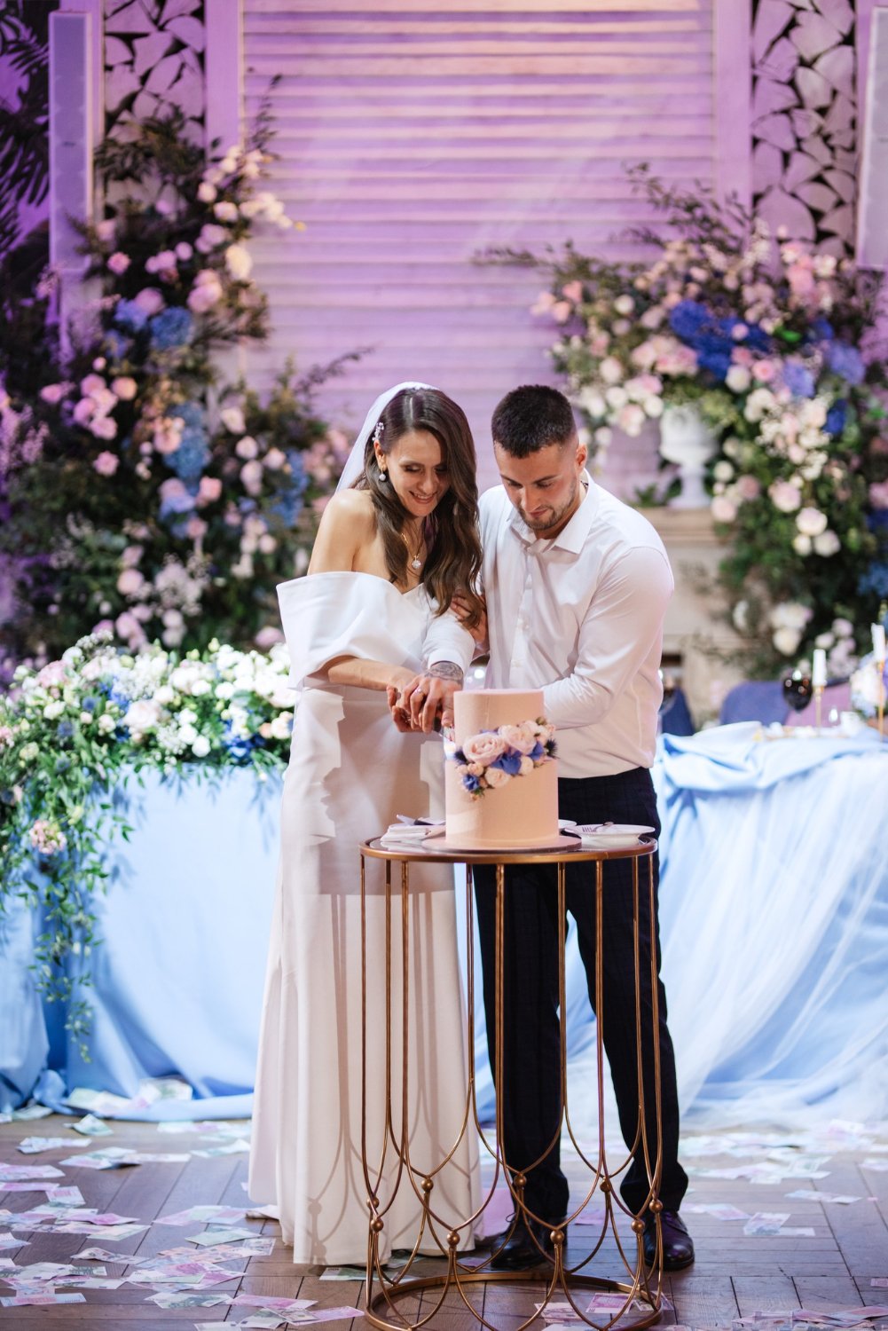 A couple dressed in wedding attire cutting a wedding cake at their reception, with floral arrangements and a decorated backdrop behind them.