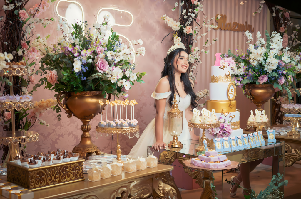 A young woman celebrating her 15th birthday, smiling and standing behind a decorated table with a large white and gold birthday cake, floral arrangements, and various desserts, in an elegant decorated room.