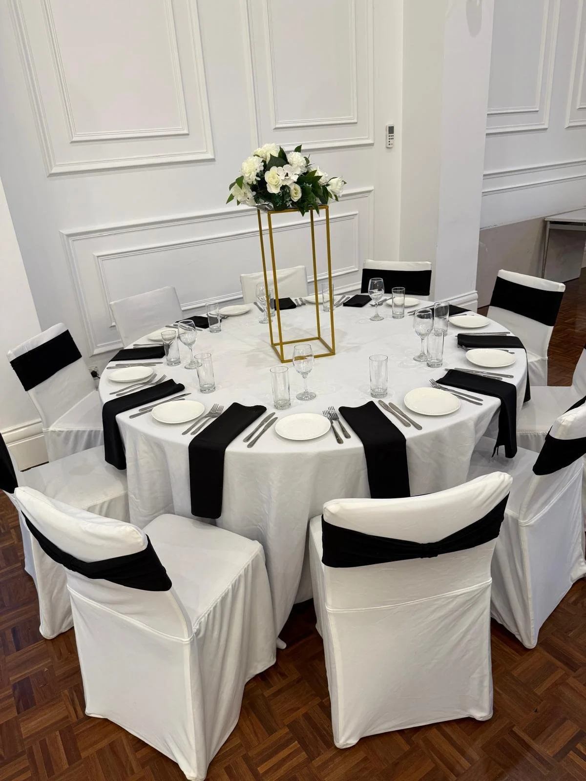 Round dining table with white tablecloth, black napkins, white chairs with black sashes, and a tall gold flower arrangement centerpiece with white flowers.
