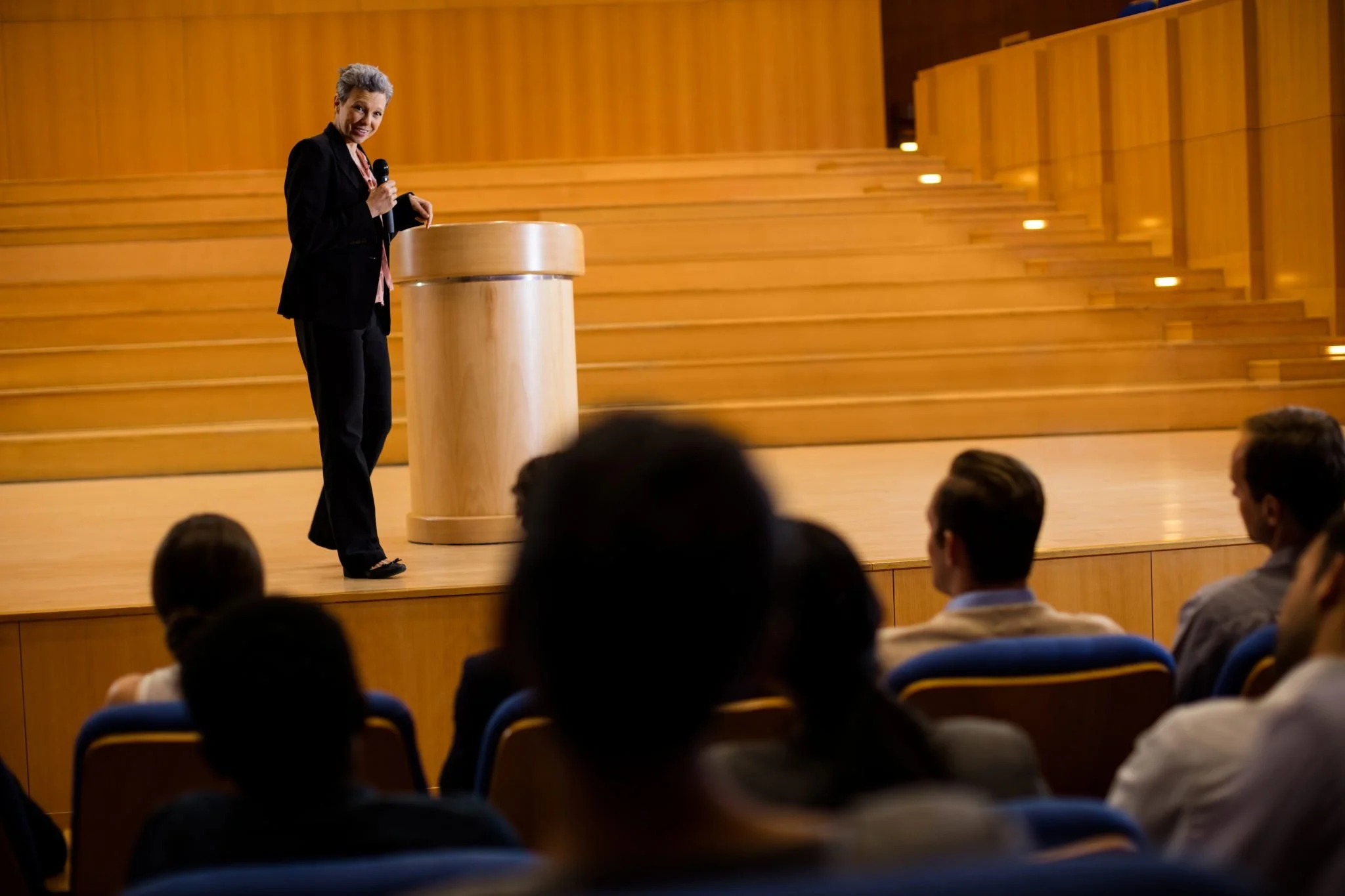 A woman standing on a stage with a wooden podium, holding a microphone, speaking to an audience in a large auditorium with wooden tiered seating.