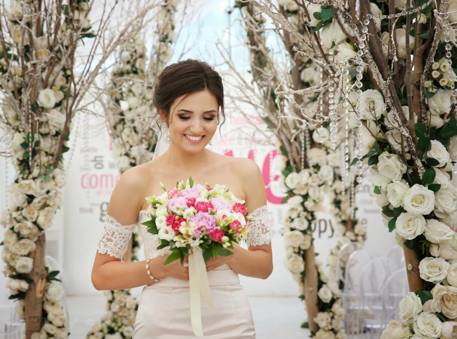 A smiling bride in a white off-shoulder wedding dress holding a pink and white bouquet, standing under a floral arch decorated with white roses and crystal beads, with a joyful expression at her wedding.