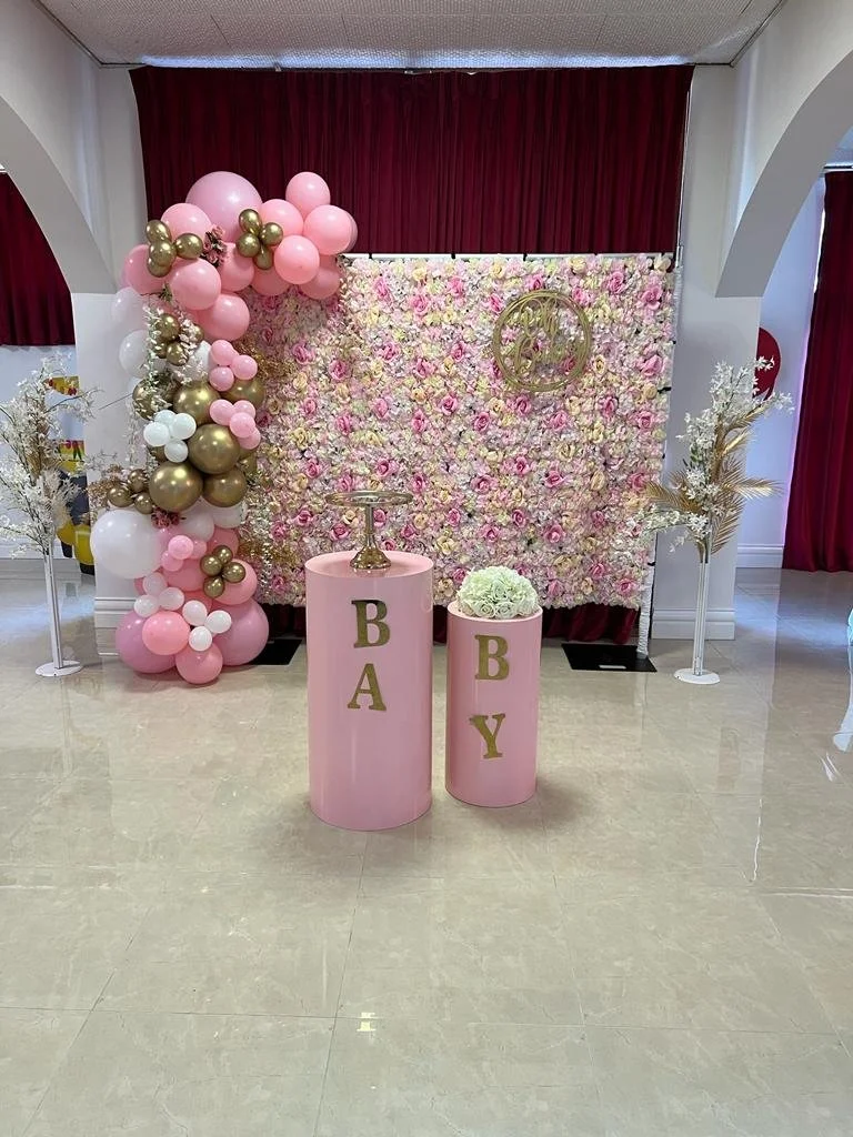 Decorative baby shower setup with pink and gold balloons, floral backdrop, and pink pillars with gold lettering 'BABY'.