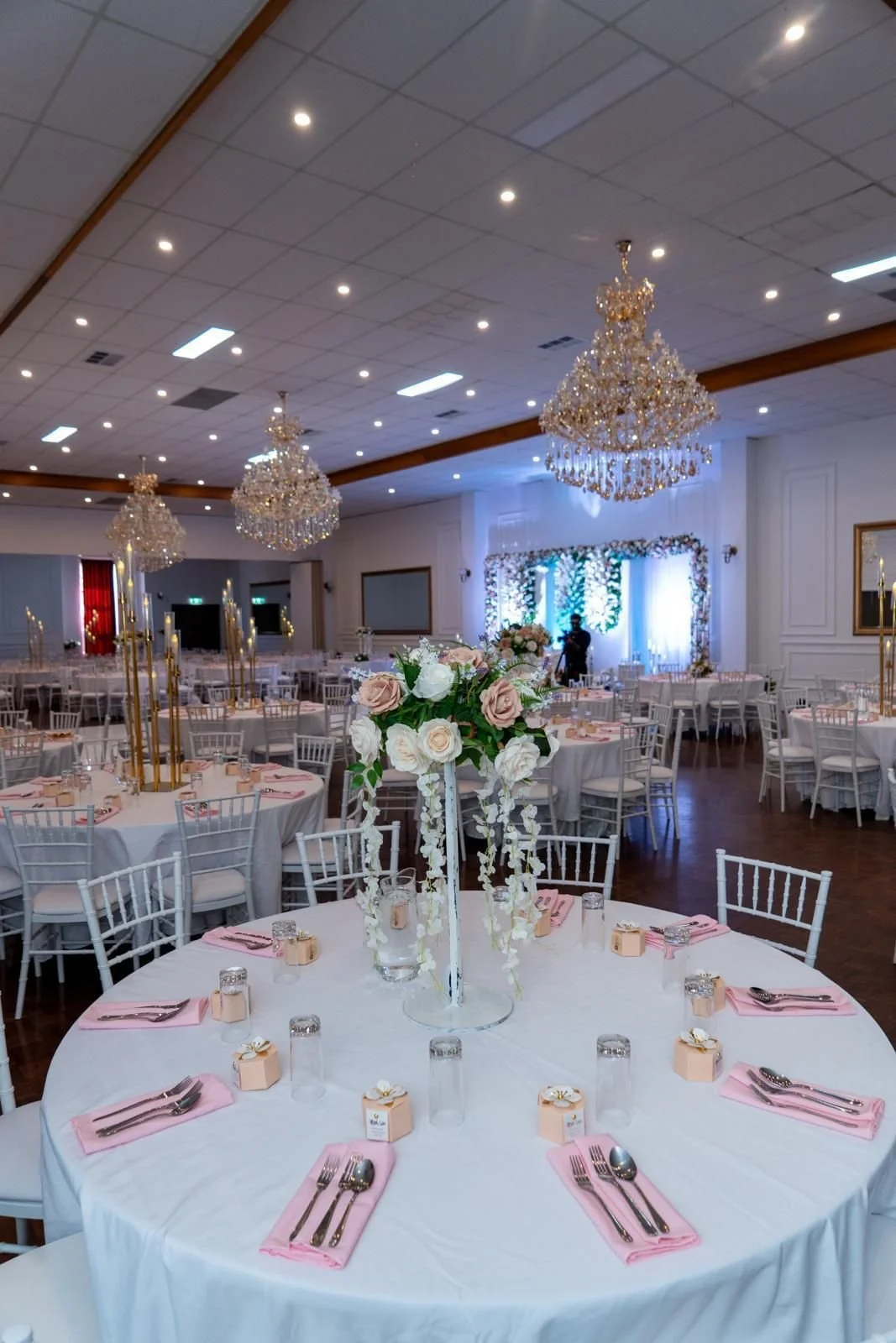 Elegant banquet hall decorated for a wedding reception with round tables covered in white tablecloths, pink napkins, and silverware, topped with a floral centerpiece of white and pink roses, and adorned with tall golden candlesticks and candles. The 
