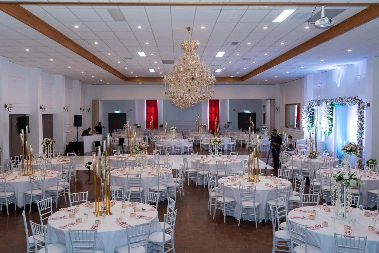 Elegant banquet hall decorated for a wedding reception with round tables, white chairs, floral centerpieces, tall gold candles, a large chandelier, and a flower arch near a side wall.