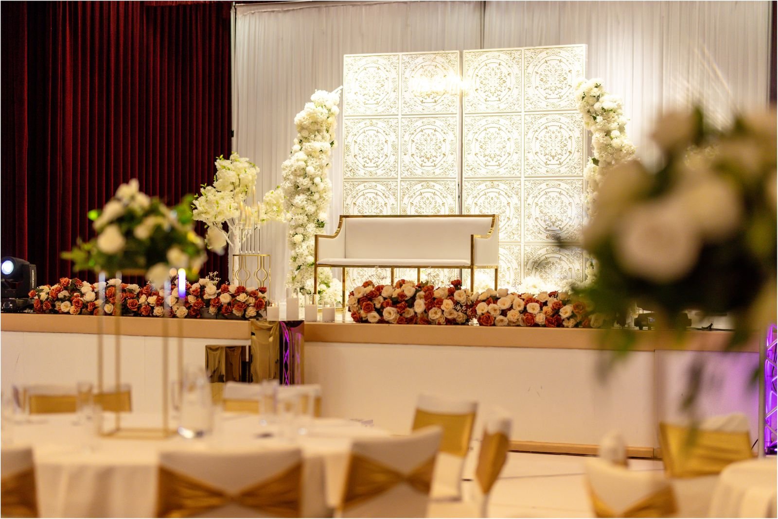 Elegant wedding stage decorated with white and red flowers, with a white bench and ornate white backdrop, set in a banquet hall with round tables and floral centerpieces.