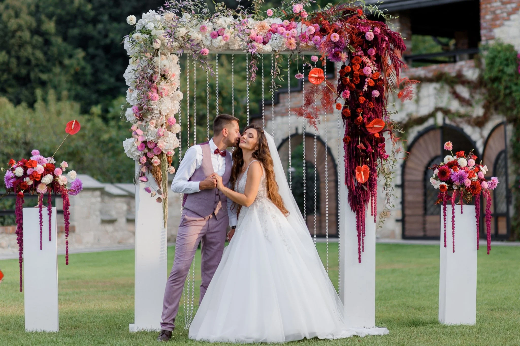 A bride and groom holding hands and about to kiss under a floral wedding arch with pink, red, and white flowers, set outdoors on green grass.