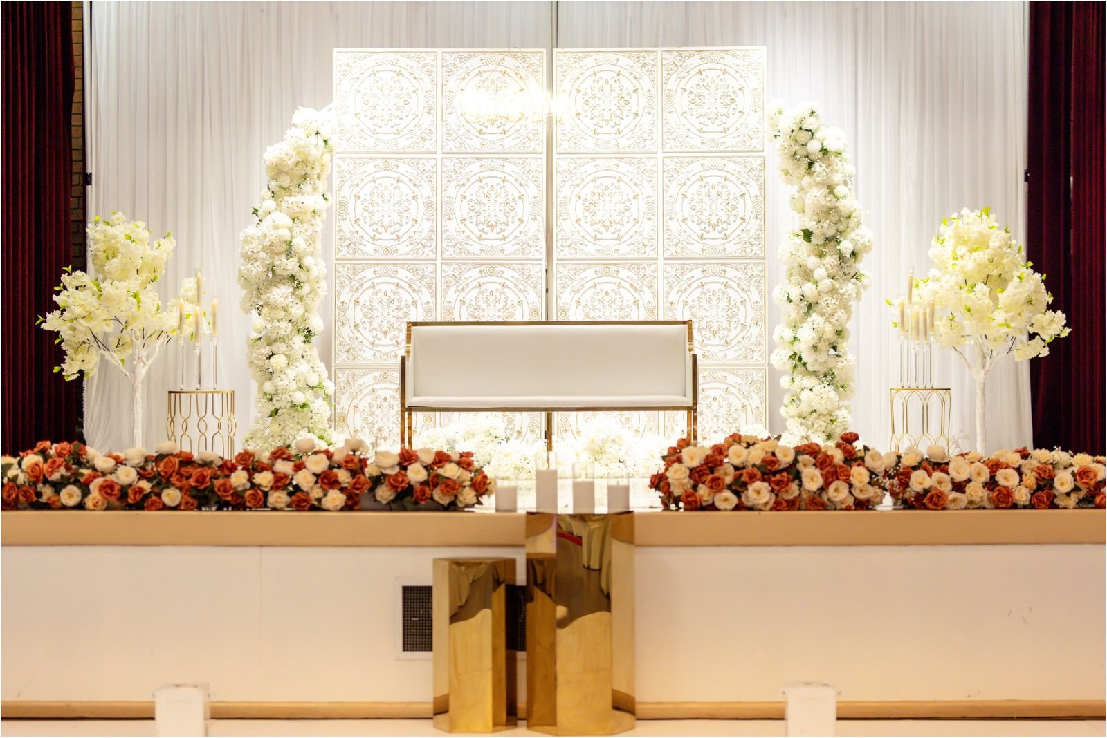 Wedding ceremony stage decorated with white and peach flowers, gold accents, and ornate backdrop panels, with a white seating bench at the center.