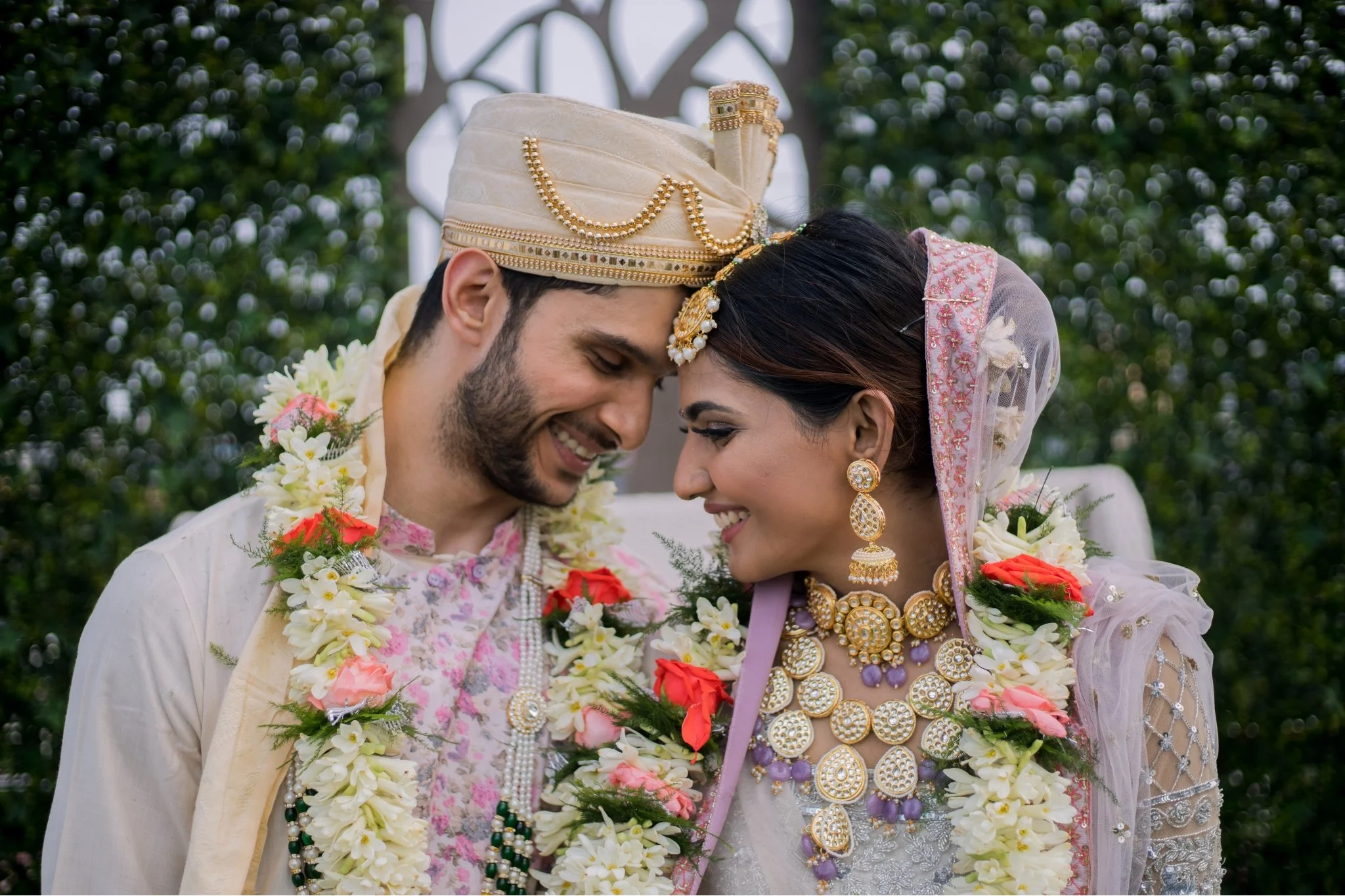 A bride and groom wearing traditional attire and jewelry, sharing a close moment during their wedding ceremony, adorned with flower garlands outdoors.