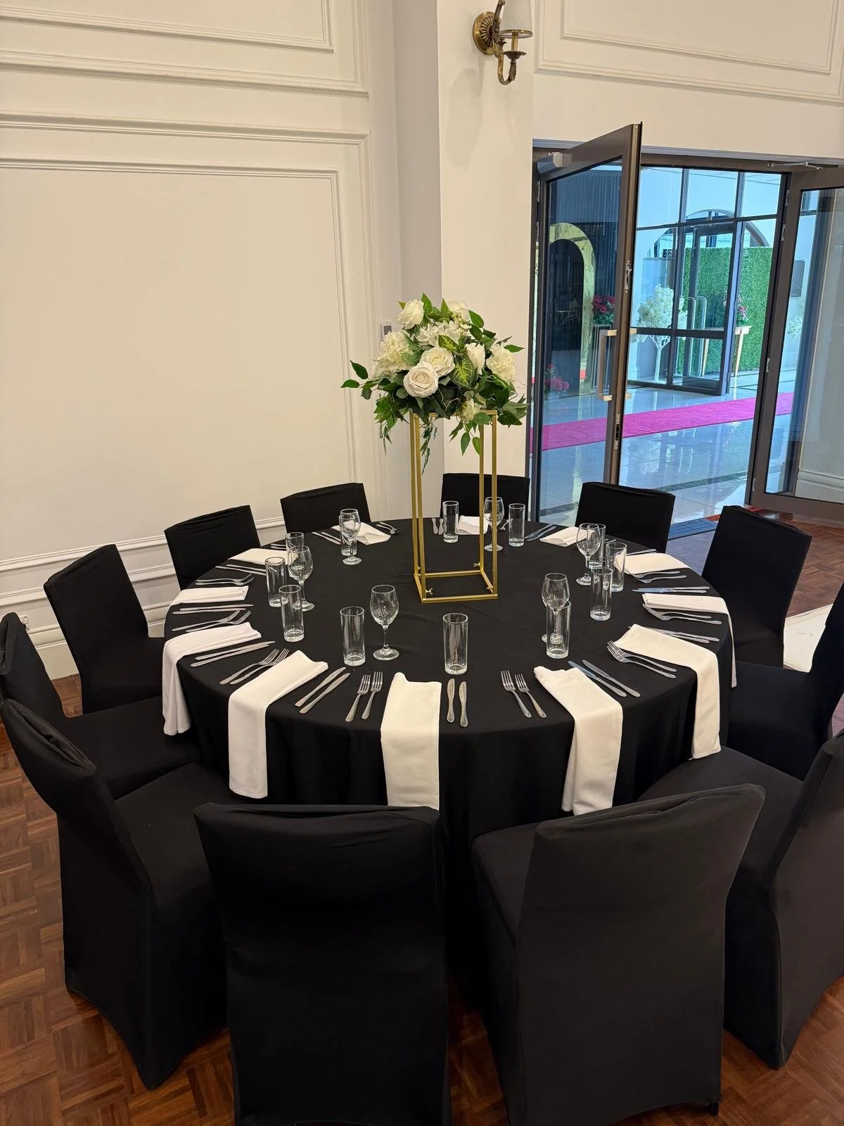 Elegant round table set with black tablecloth, white cloth napkins, silverware, and glassware. A tall gold geometric stand holds a lush floral centerpiece of white roses and greenery. The table is in a room with white walls, a partially open glass do