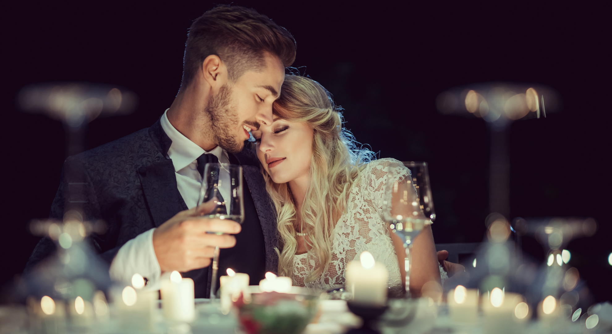 A romantic moment at a dinner date with a man and woman with their foreheads touching, surrounded by candles and glasses of wine.