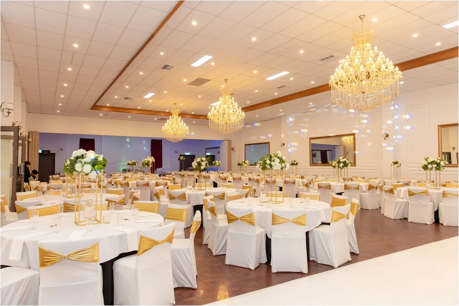 Elegant banquet hall decorated for a formal event with white tablecloths, gold bows on chairs, and tall floral centerpieces. The room features three large chandeliers hanging from a white ceiling with recessed lighting, and dark framed mirrors on the