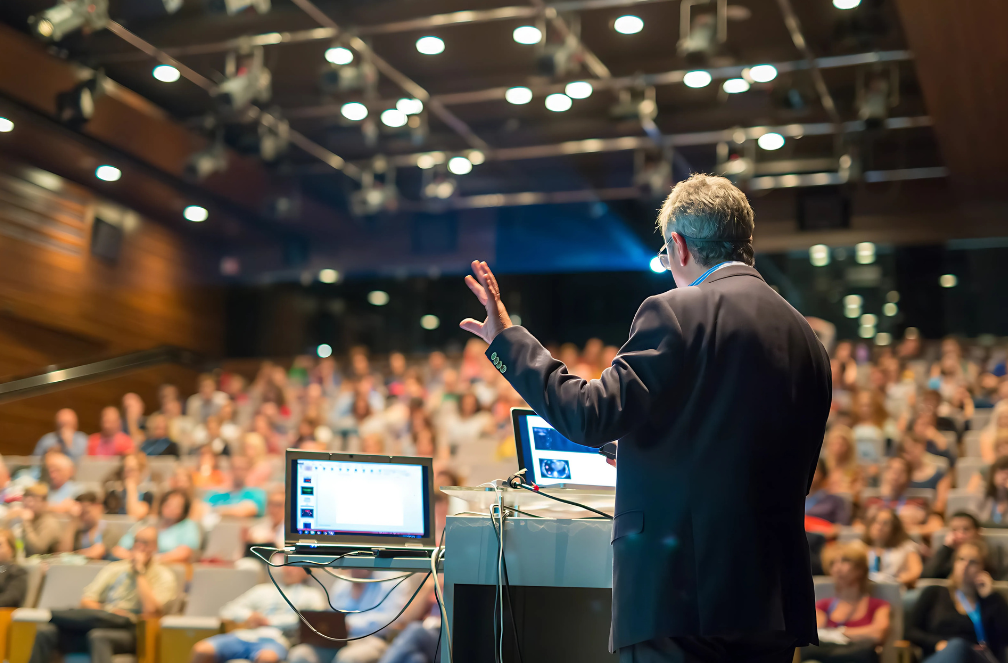 A man in a suit presenting at a conference with an audience in a large auditorium.