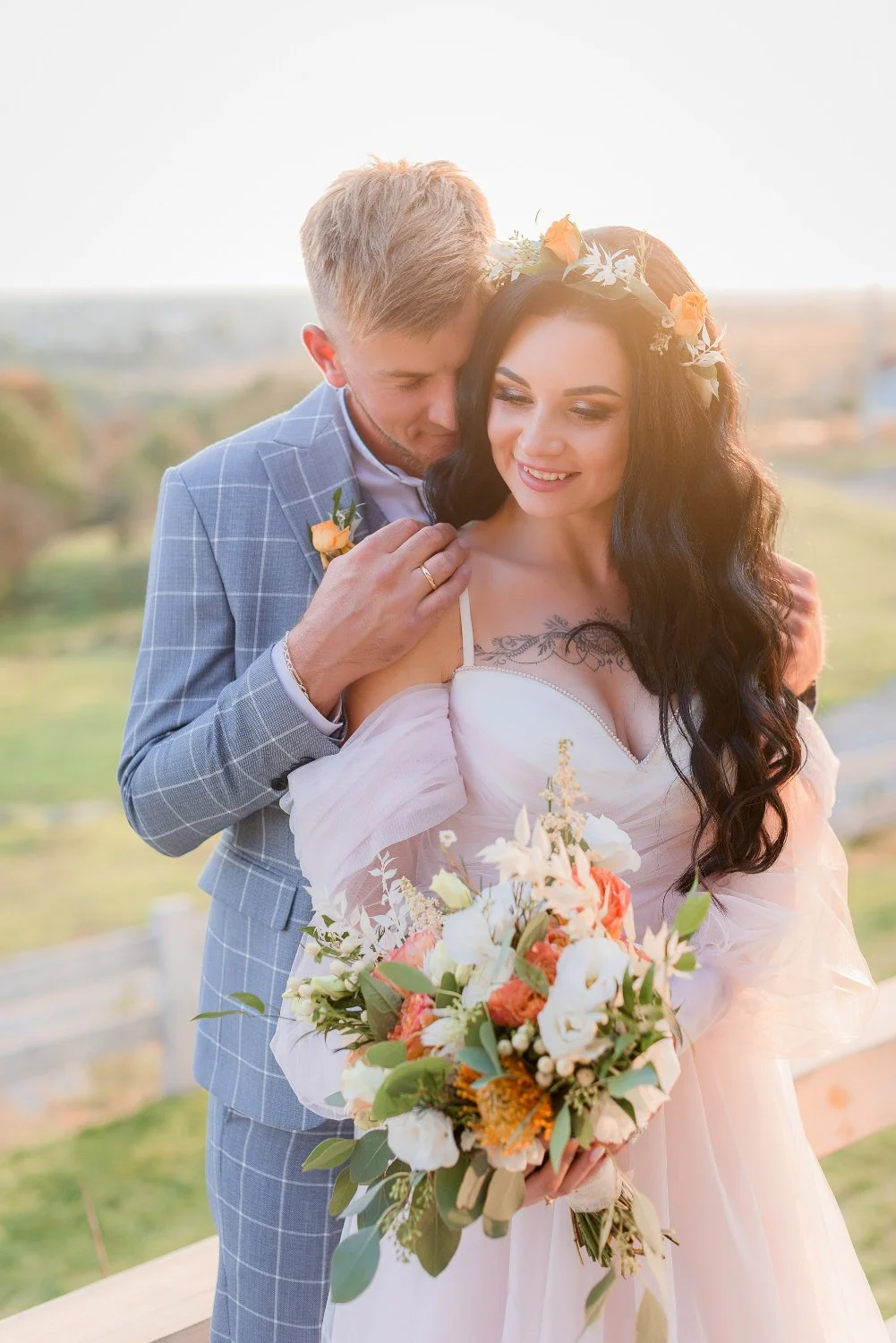 A newlywed couple sharing a romantic moment outdoors during sunset, with the groom embracing the bride. The bride holds a colorful bouquet of flowers and wears a floral crown, while the groom is dressed in a light grey suit with a small flower boutonniere.