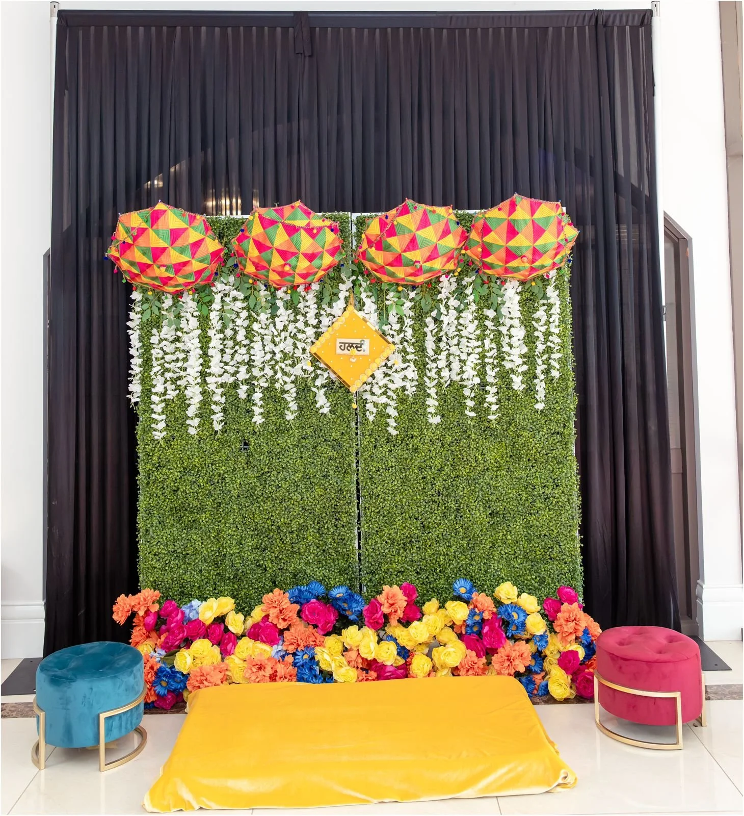 Decorative backdrop with black curtains, green leafy wall, white flowers, colorful umbrellas, a yellow sign with 'शुभ', and vibrant flower arrangements at the base, with blue and pink velvet stools.