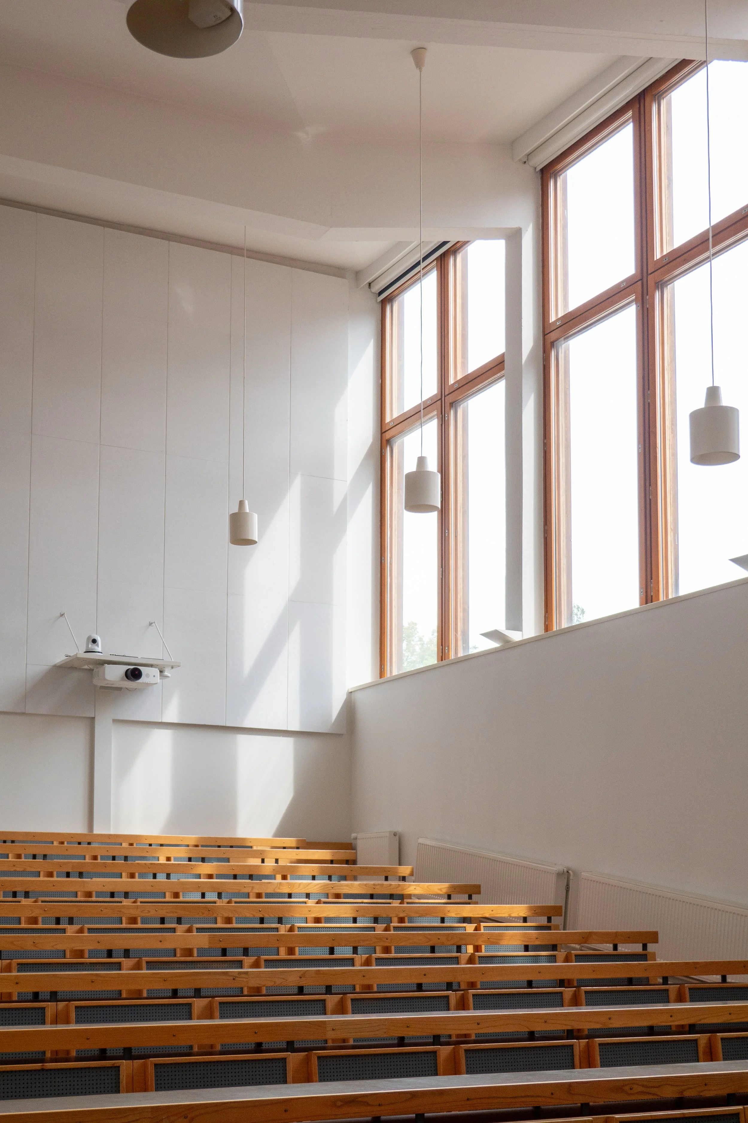Empty lecture hall with wooden benches, large windows letting in sunlight, hanging white pendant lights, and a projector on the wall.
