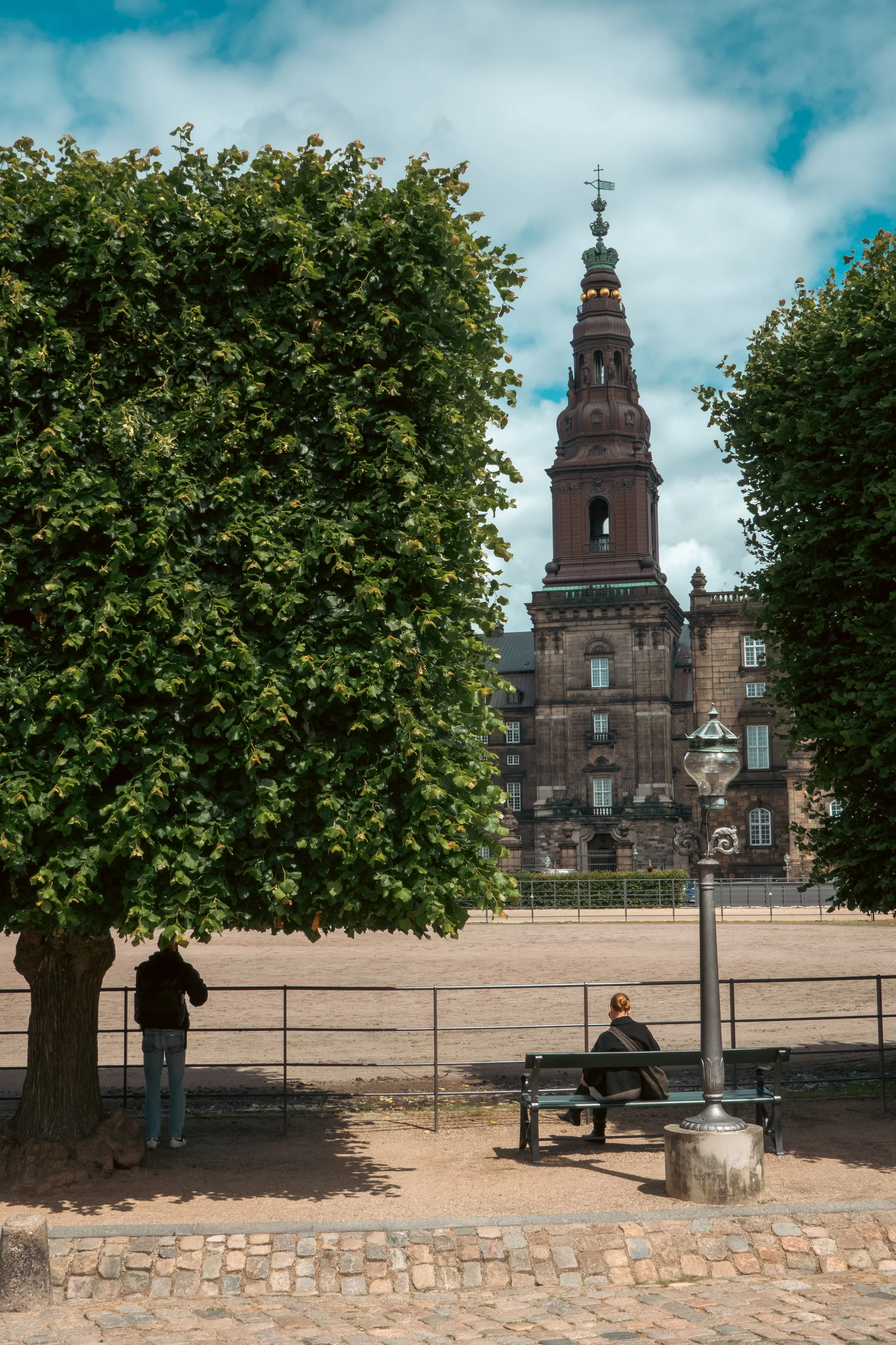 A historic church with a tall steeple seen between two green trees. Two people are sitting on a park bench, and another person stands near a fence, with a lamppost nearby.