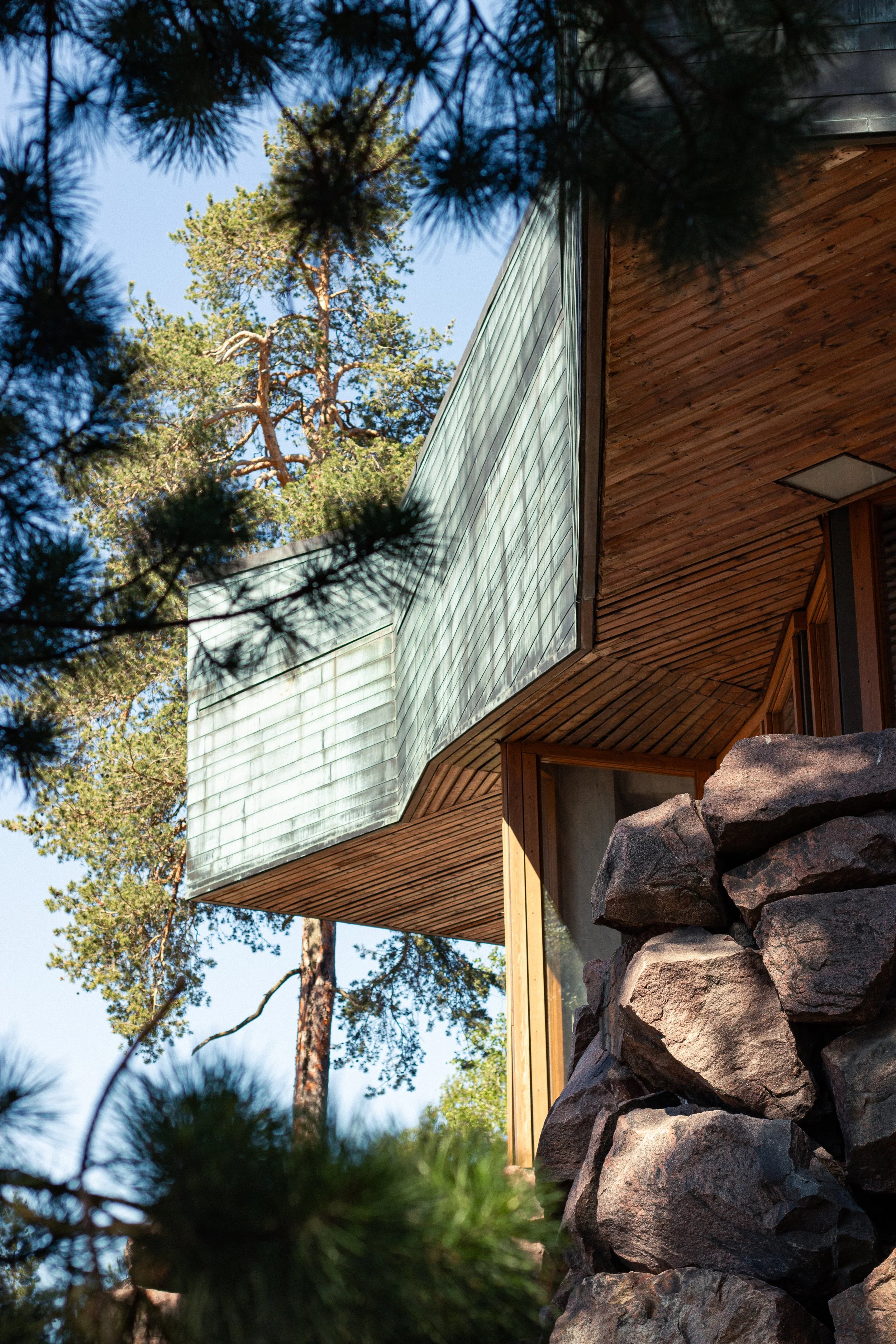 Modern house with wooden exterior, large rock foundation, green metal roof, surrounded by pine trees and clear blue sky.