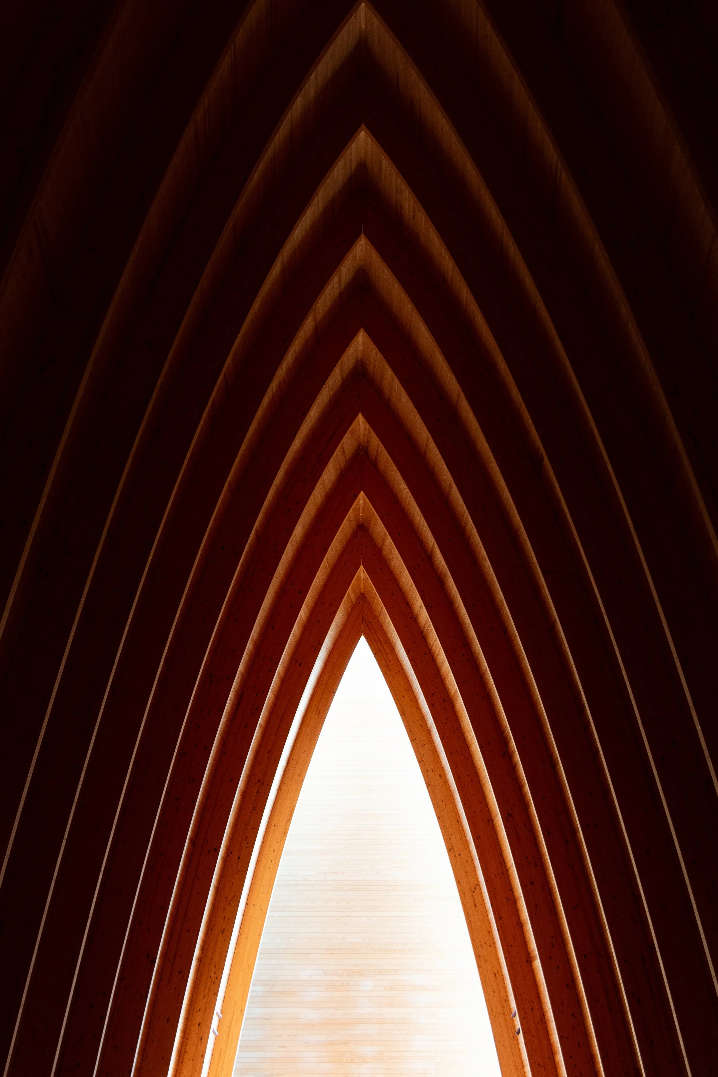 Interior view of wooden architectural structure with arched beams pointing toward a bright light at the top.